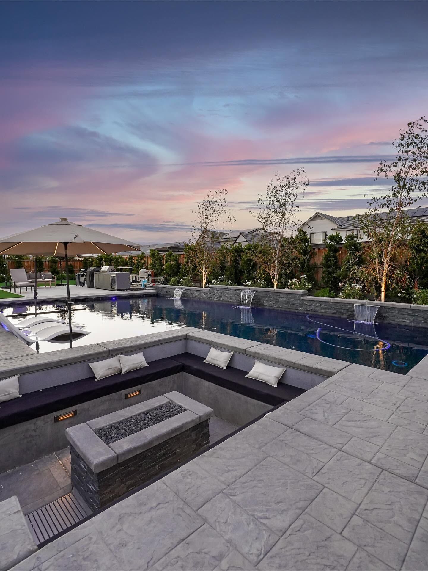 Modern backyard pool and fire pit at sunset with blue water, patio seating, and pink sky