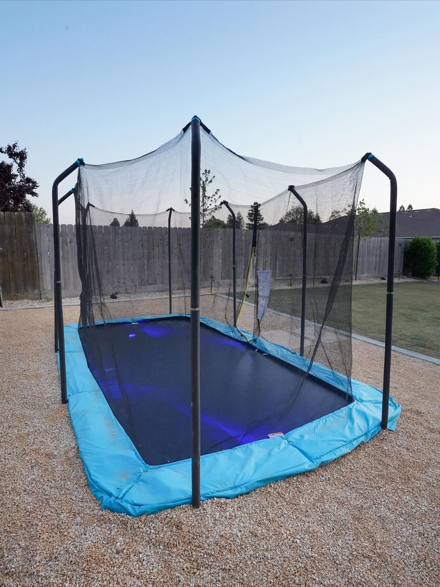 Black trampoline net enclosure with blue padding on a gravel yard under a clear sky