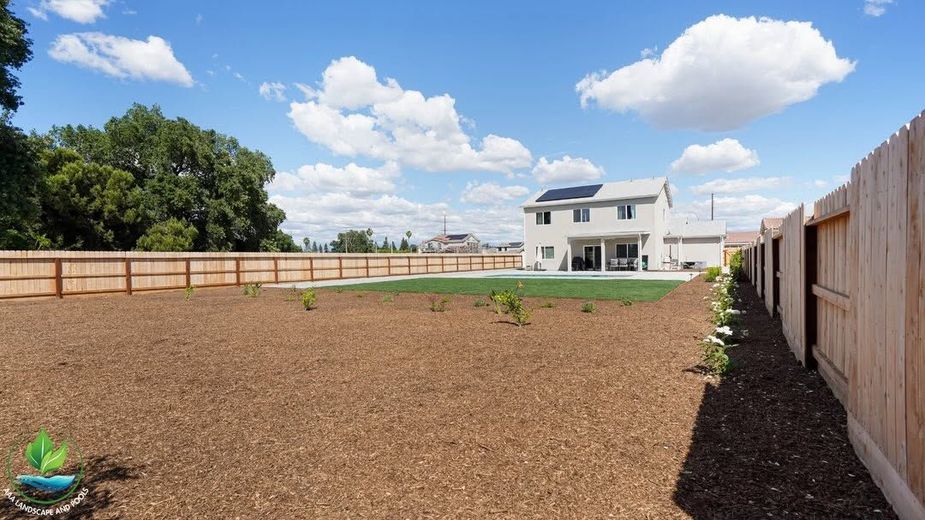 Freshly mulched backyard with a white house, wooden fences, and blue sky with clouds