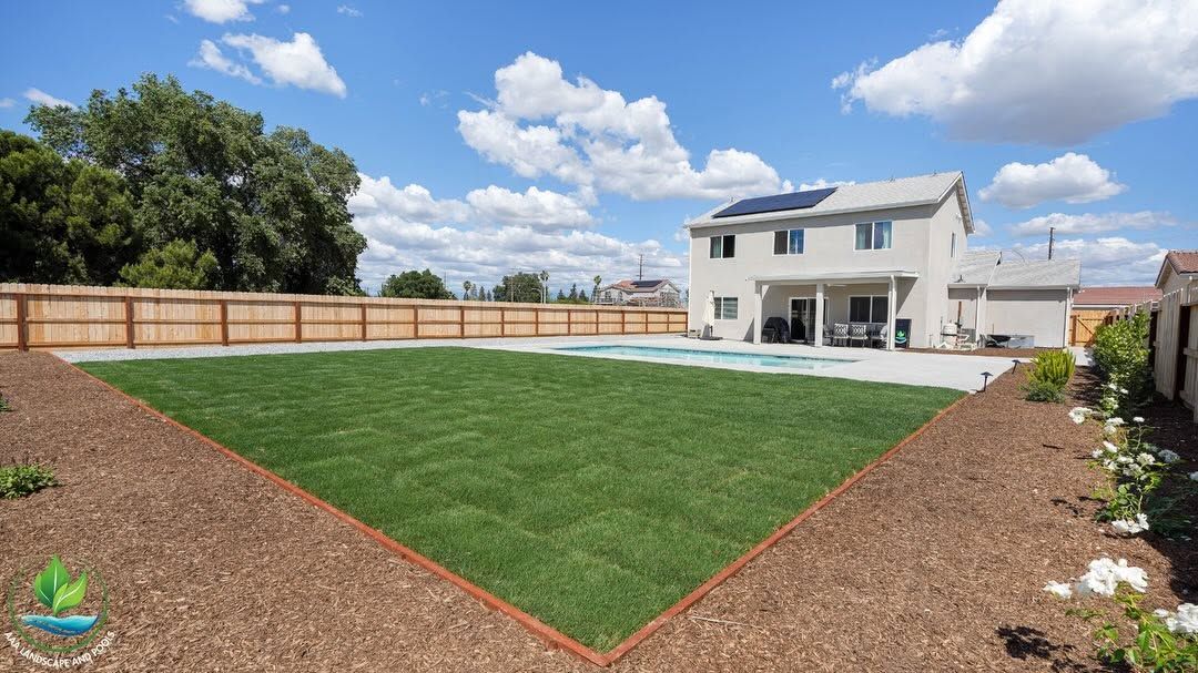 Backyard with green lawn, mulch borders, wooden fence, and a white two-story house under a blue sky