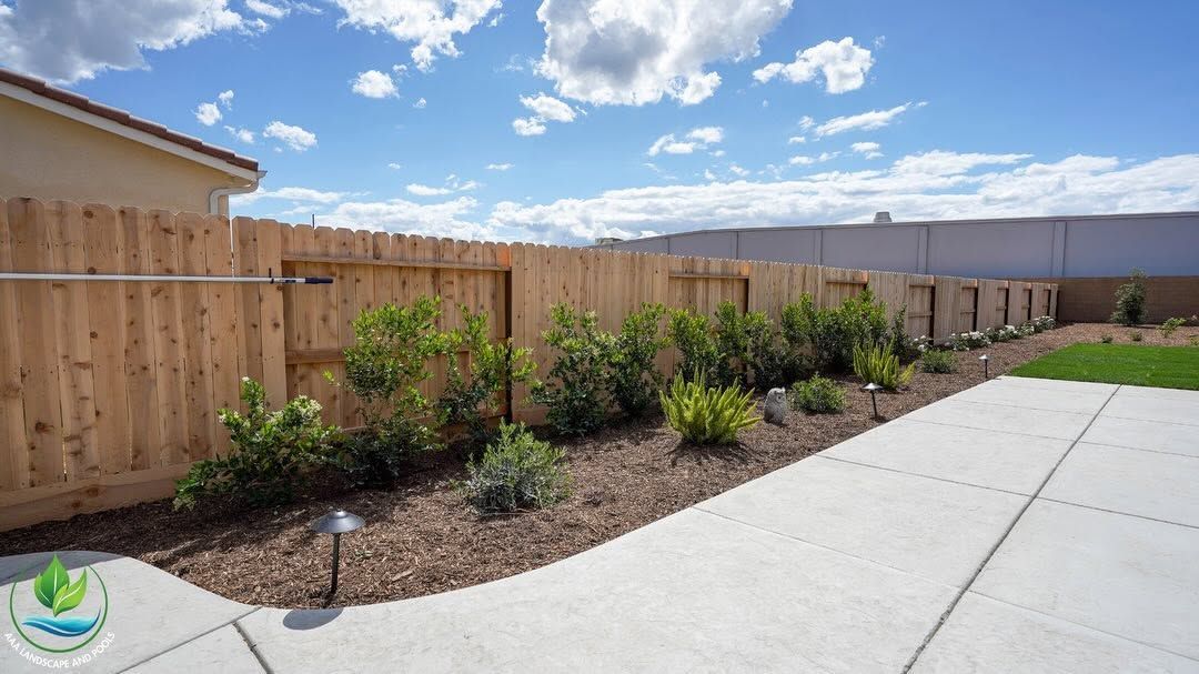 Newly landscaped backyard with wooden fence, shrubs, and a concrete patio under a blue sky