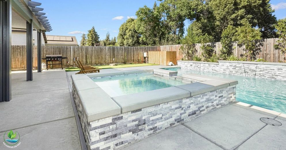 Backyard pool with raised spa, stone deck, fence, and trees in bright sunlight