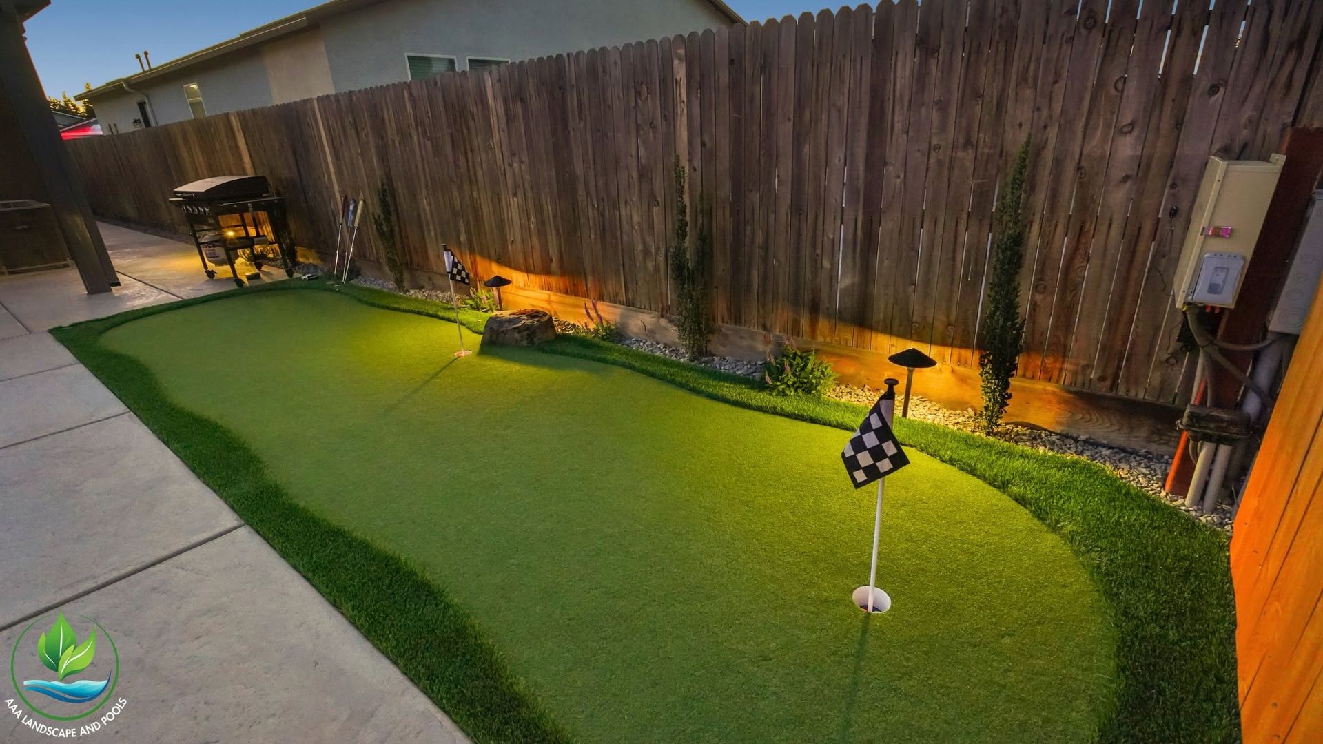 Backyard patio with artificial grass, wooden fence, warm lights, and a small garden flag.