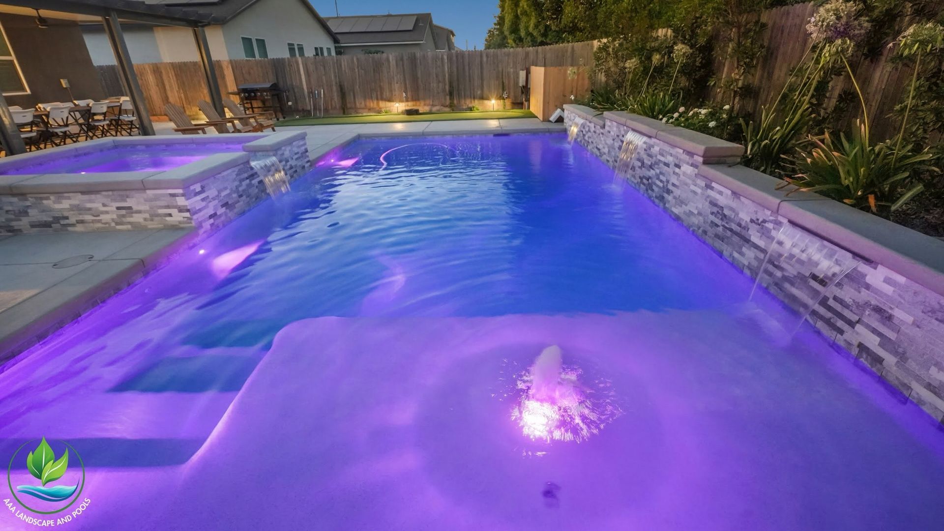 Purple-lit backyard swimming pool at dusk with glowing water and patio seating around it