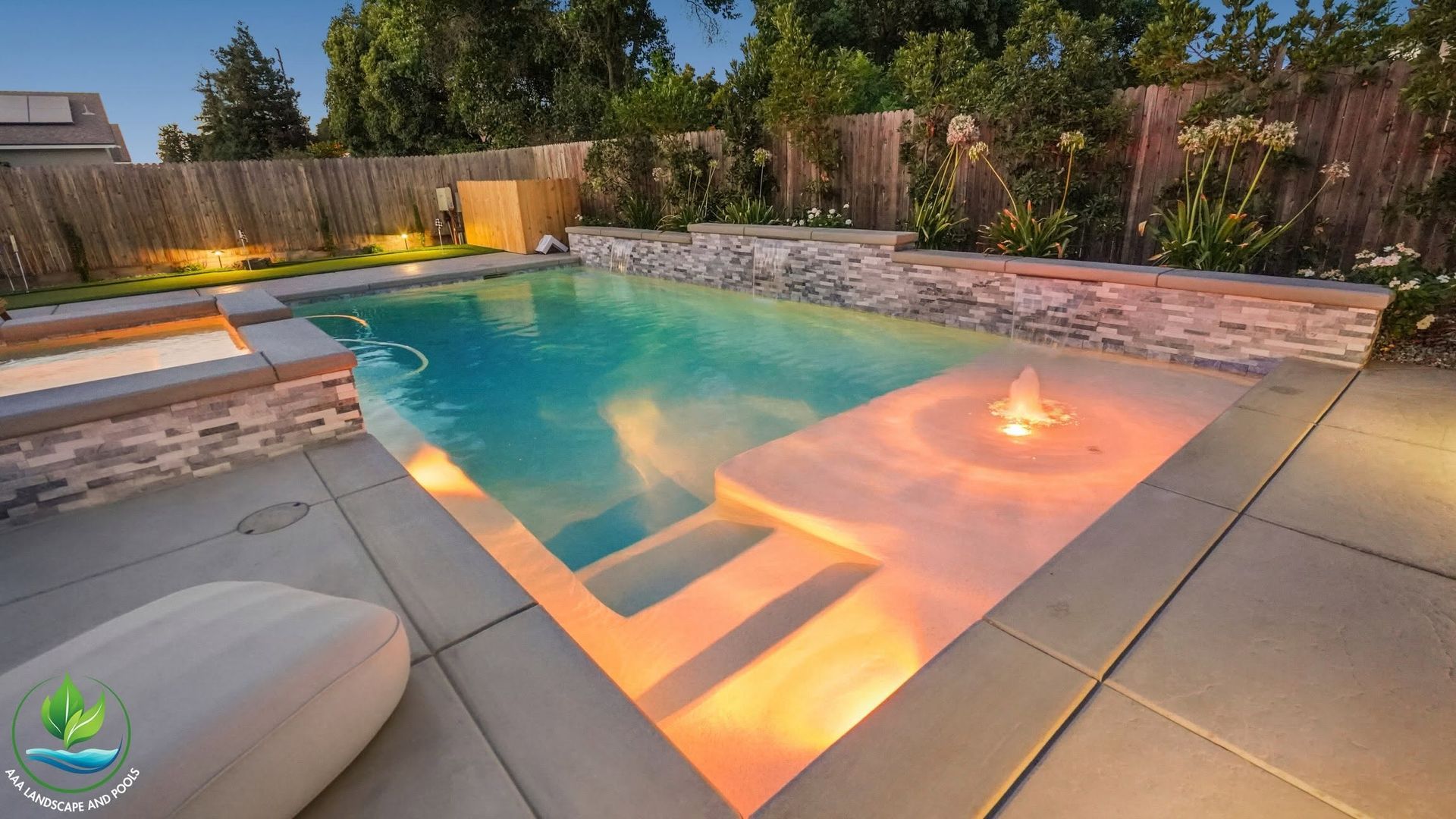 Modern backyard pool with warm lighting, fire feature, stone deck, and surrounding privacy fence at dusk