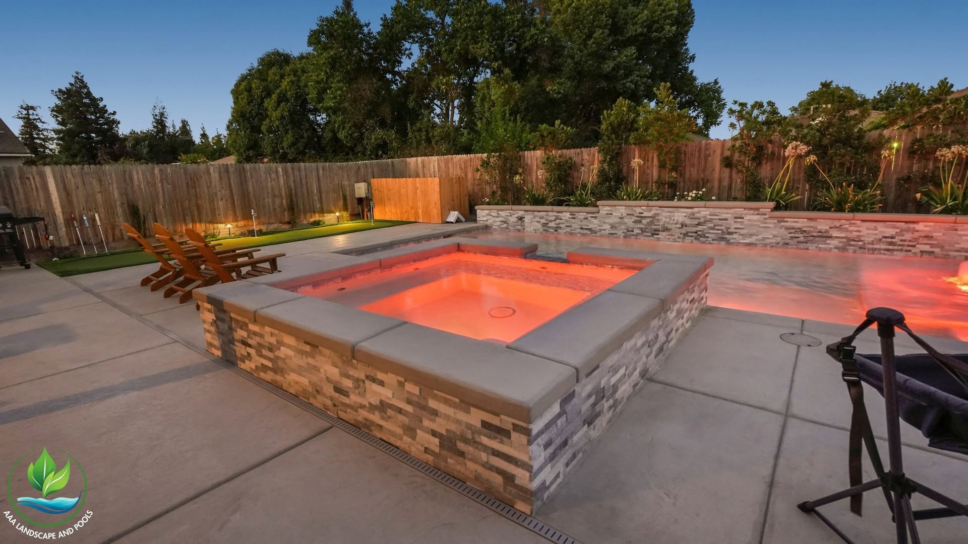 Backyard patio with a square stone fire pit glowing orange at dusk, surrounded by concrete and trees