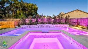 Purple-lit backyard pool with a spa, patio, and fence at dusk