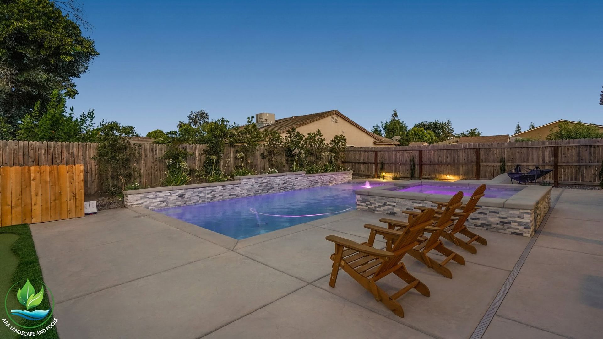 Backyard pool with lounge chairs, concrete patio, wooden fence, and hedge at dusk
