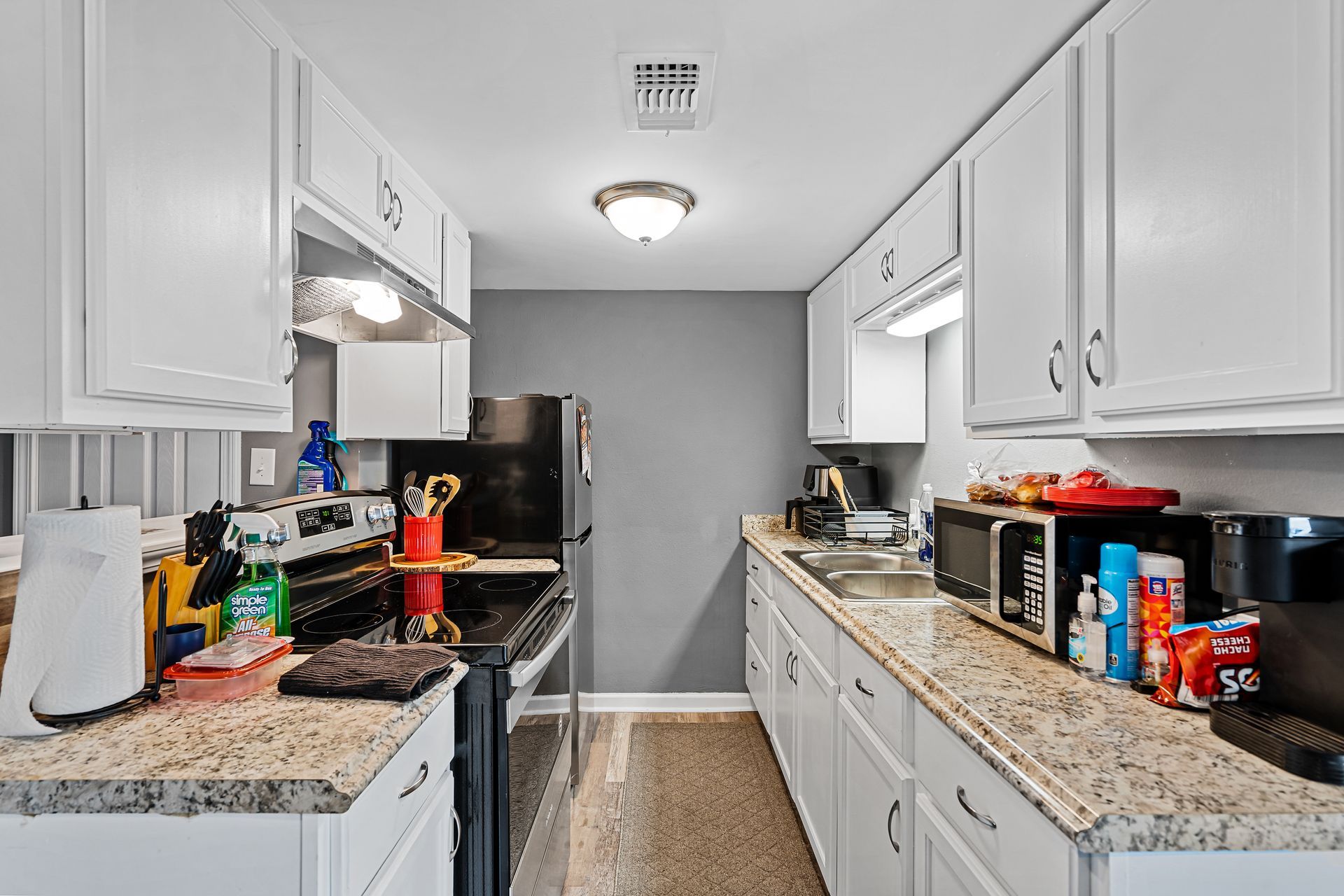 Narrow white galley kitchen with granite countertops, black appliances, and cluttered items on both sides.