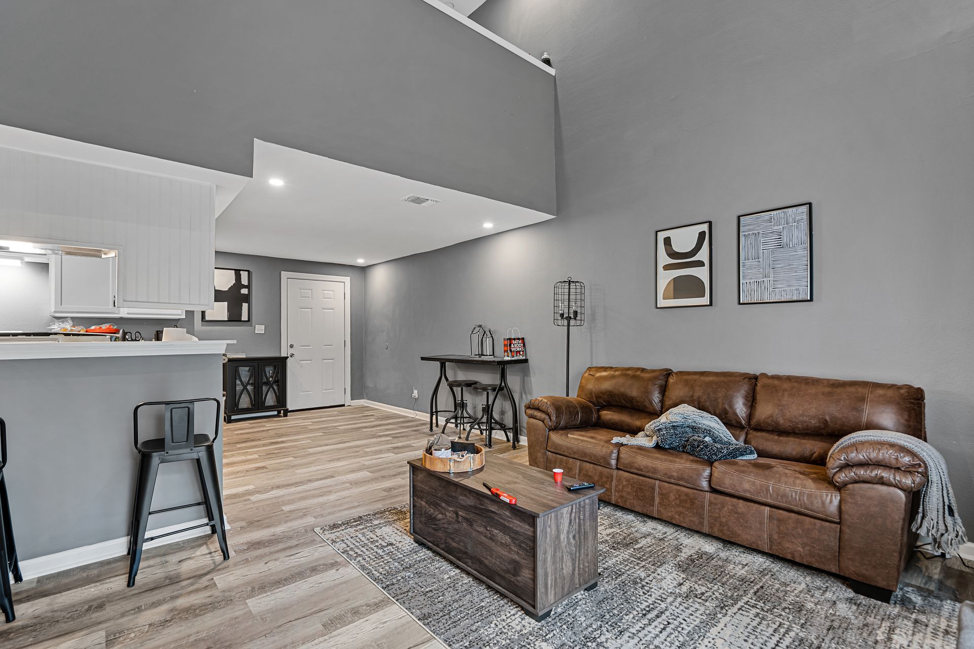 Open-plan living room with brown leather sofa, coffee table, rug, and adjoining kitchen with bar stools.
