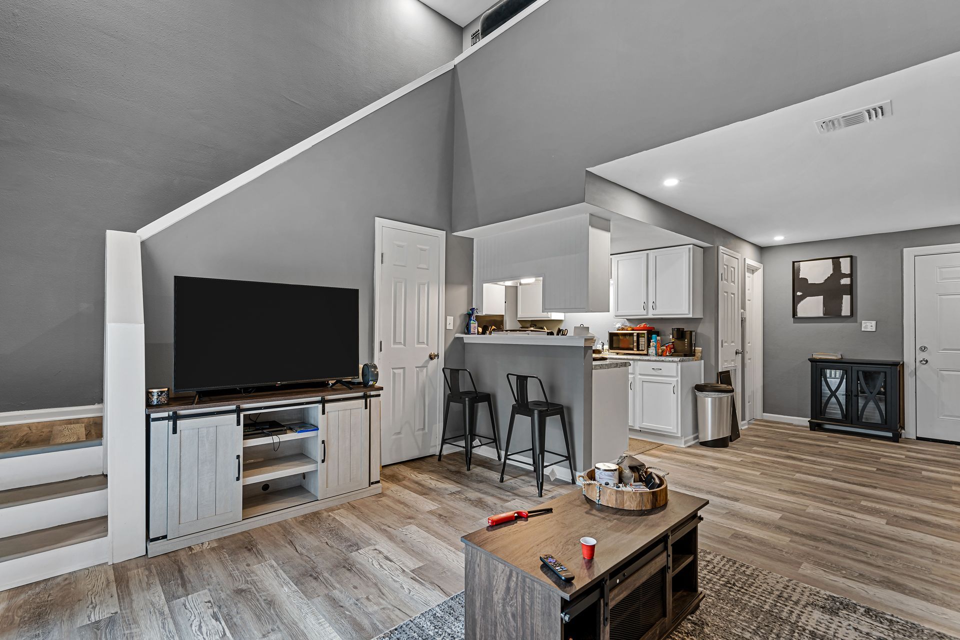 Modern open-concept living room with gray walls, TV, staircase, bar stools, and kitchen in the background