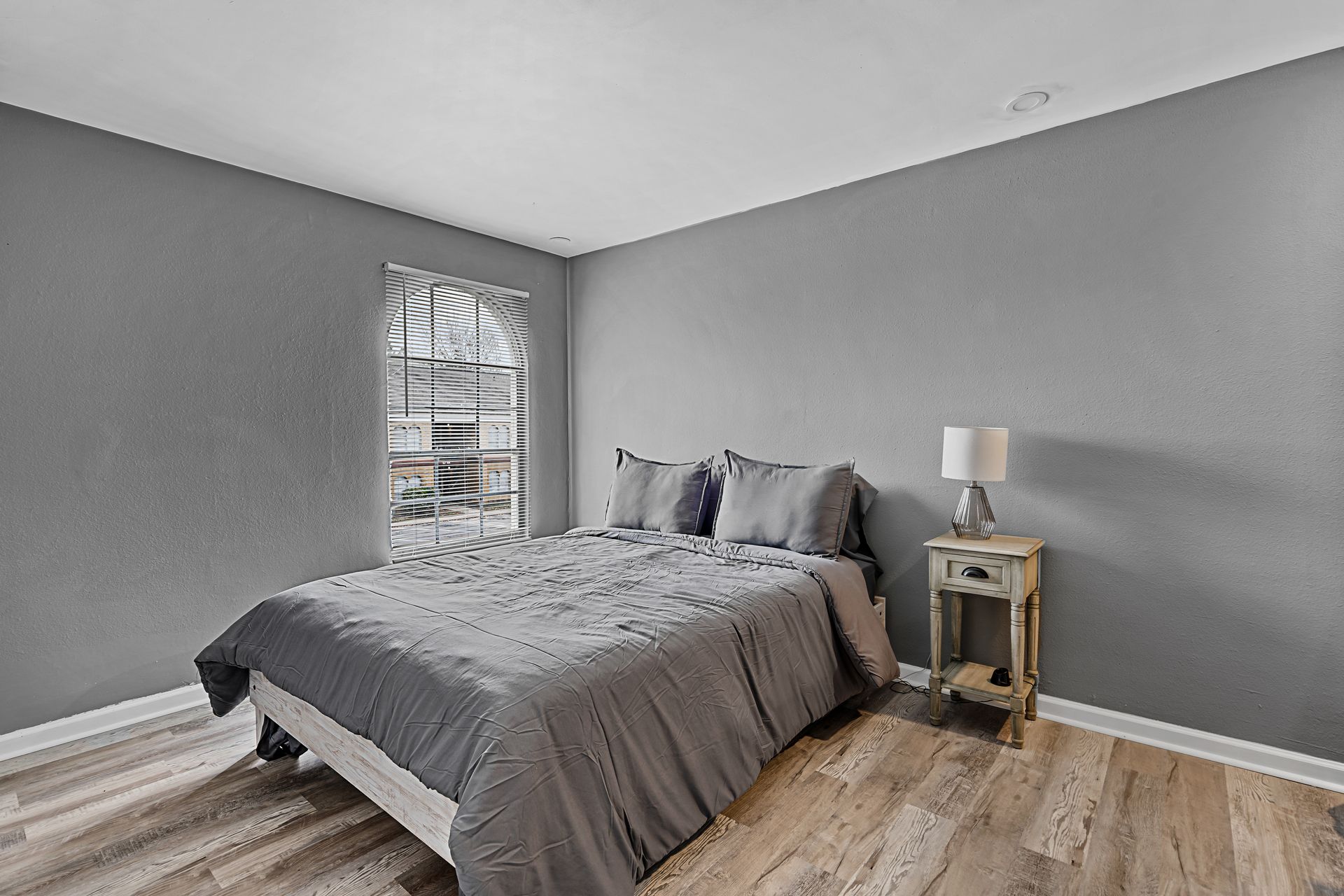 Gray bedroom with bed, nightstand, lamp, and window blinds on wood floor