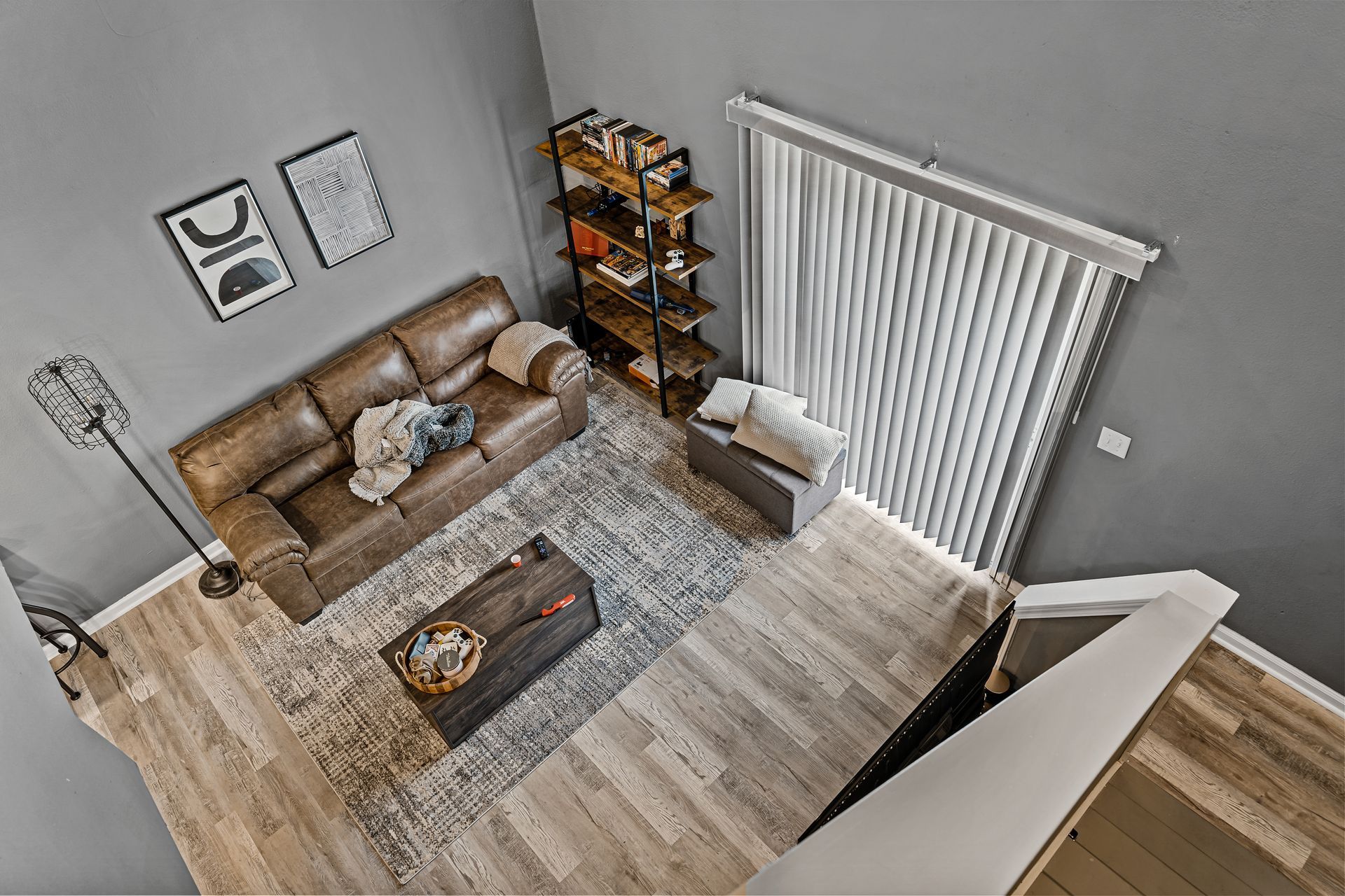 Overhead view of a cozy living room with a brown sofa, rug, coffee table, shelving, and vertical blinds.