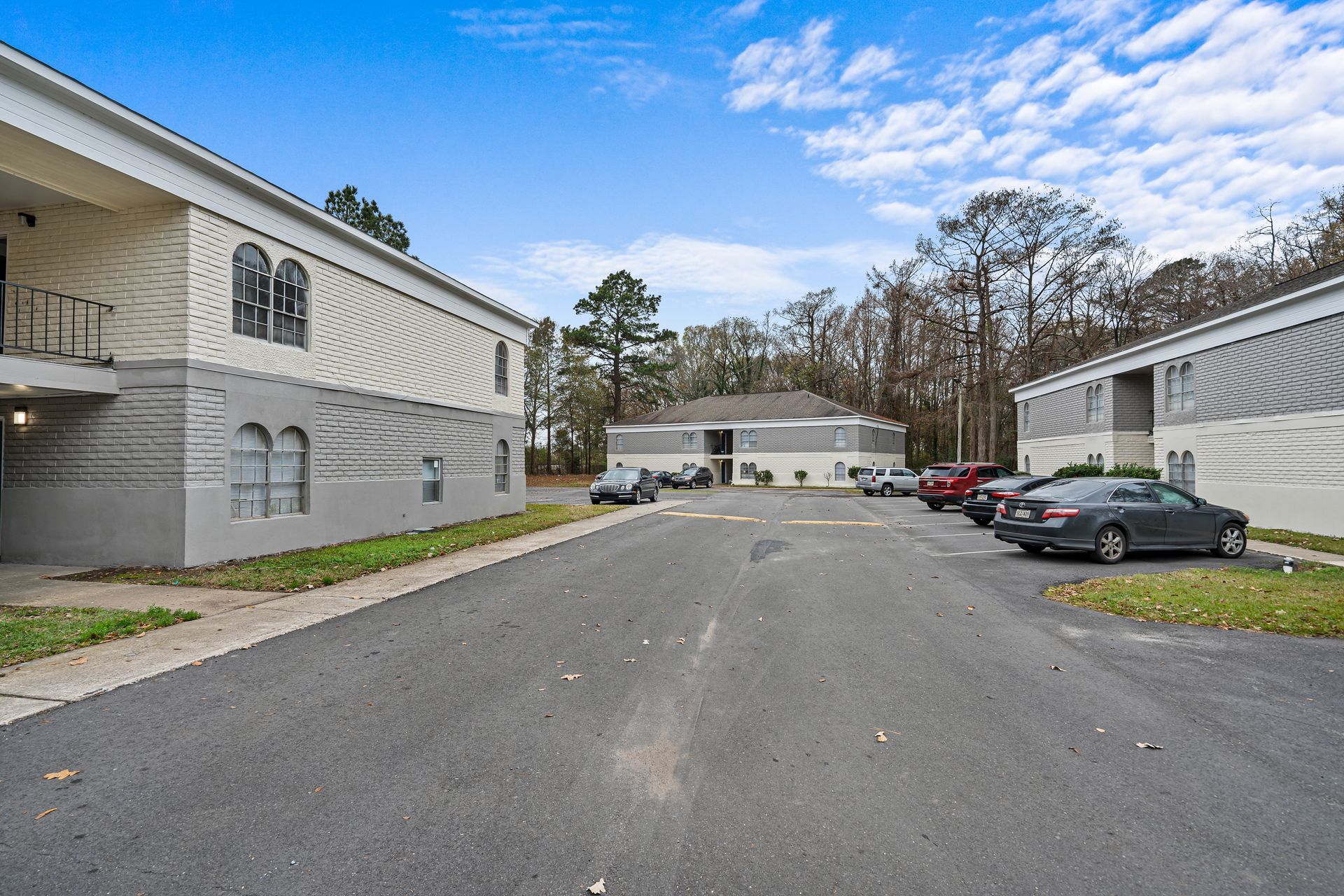 Apartment complex parking lot with cars, gray buildings, and trees under a blue sky