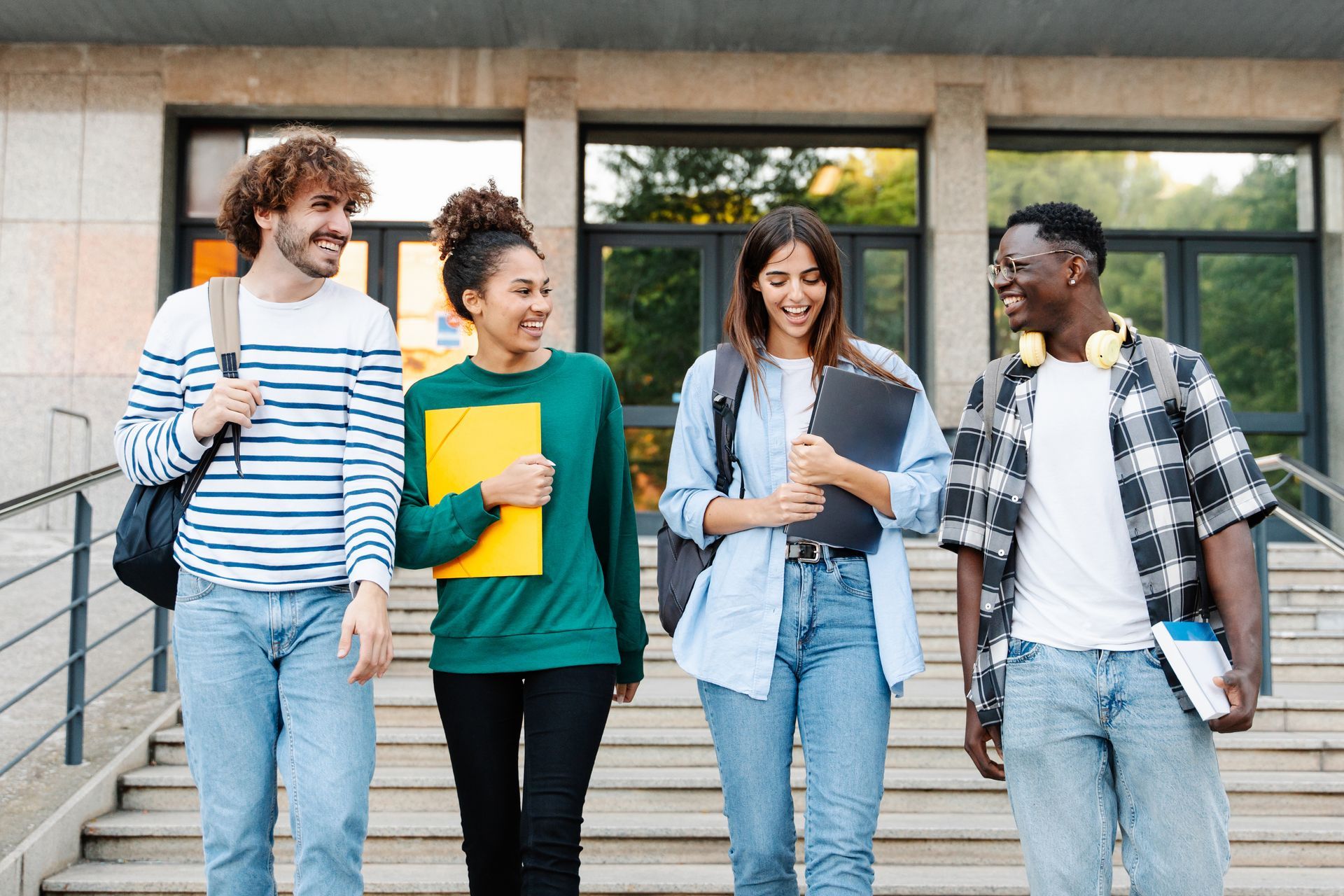 Four students walking outside a campus building, smiling and carrying notebooks and backpacks.