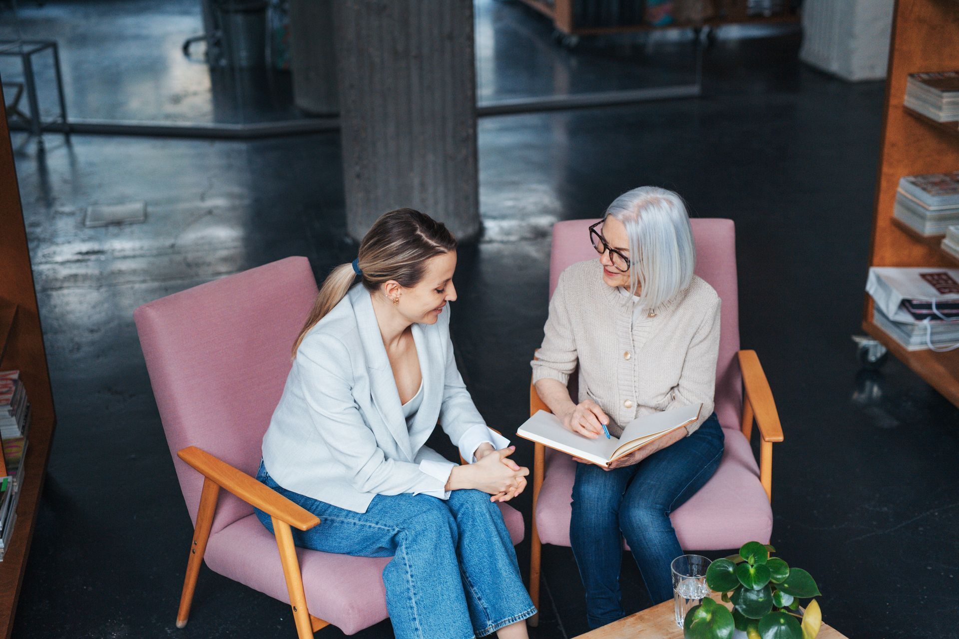 Two people seated on pink chairs, talking and looking at a notebook in a modern indoor lounge.
