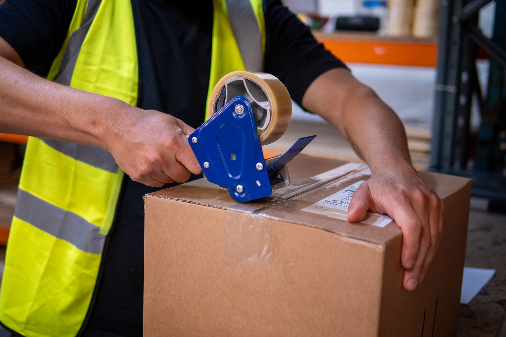 Person in a yellow vest seals a cardboard box with tape dispenser in a warehouse.