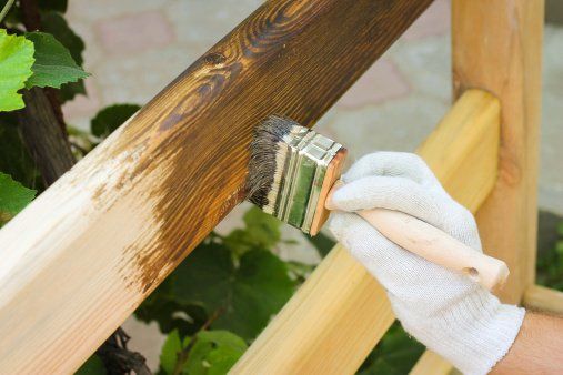 A person wearing gloves is applying wood stain to a piece of wood using a brush.