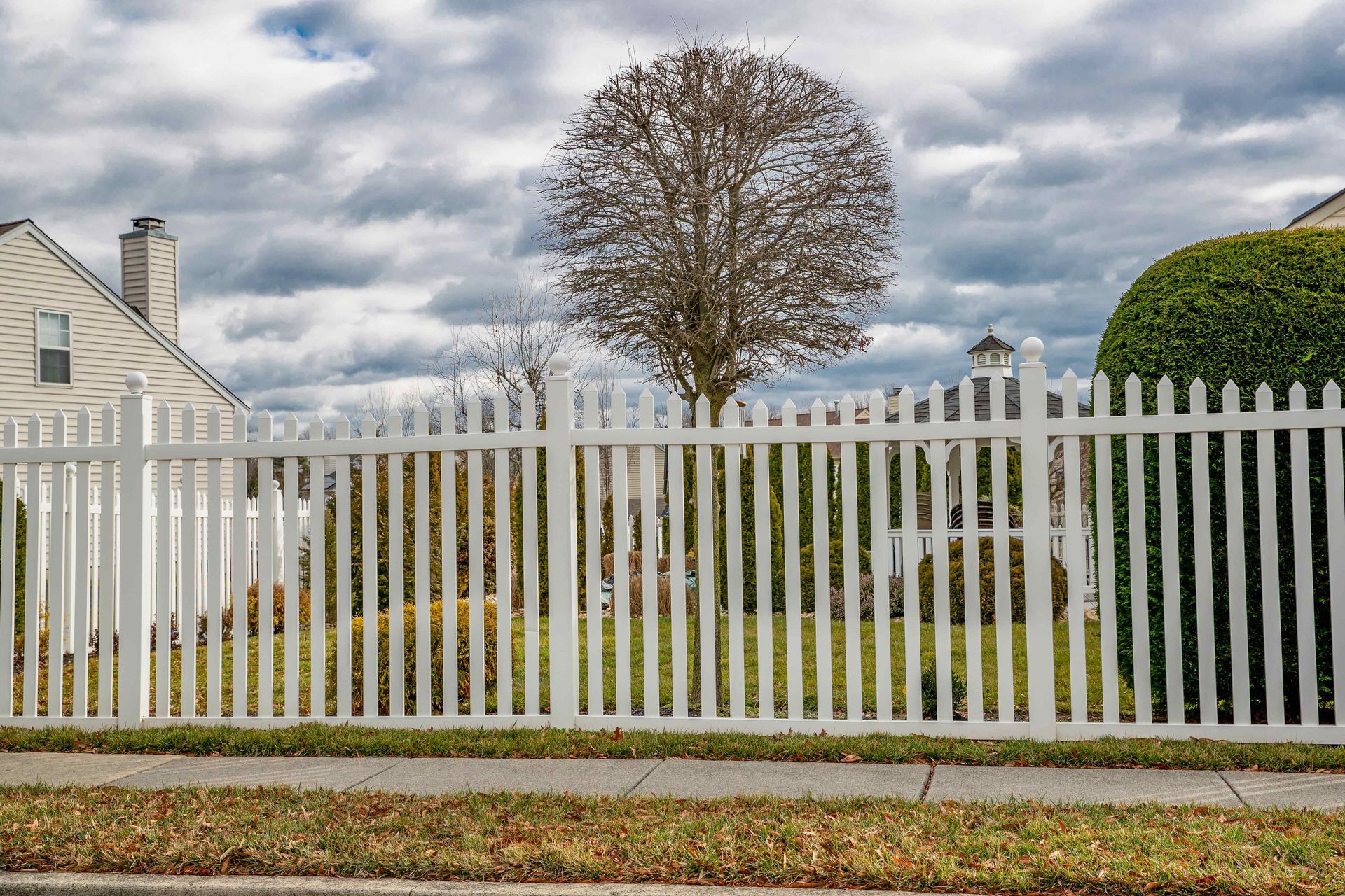 A white picket fence surrounds a house with a tree in the background.