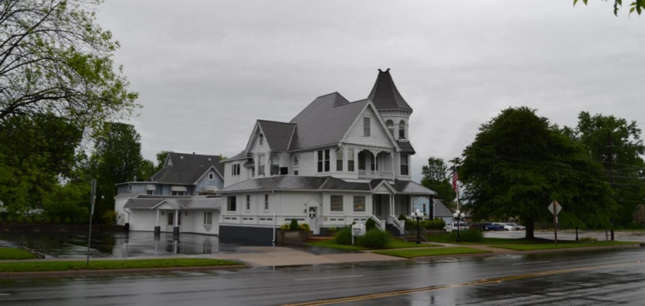 Large white Victorian house on a rainy street, with trees and overcast sky.
