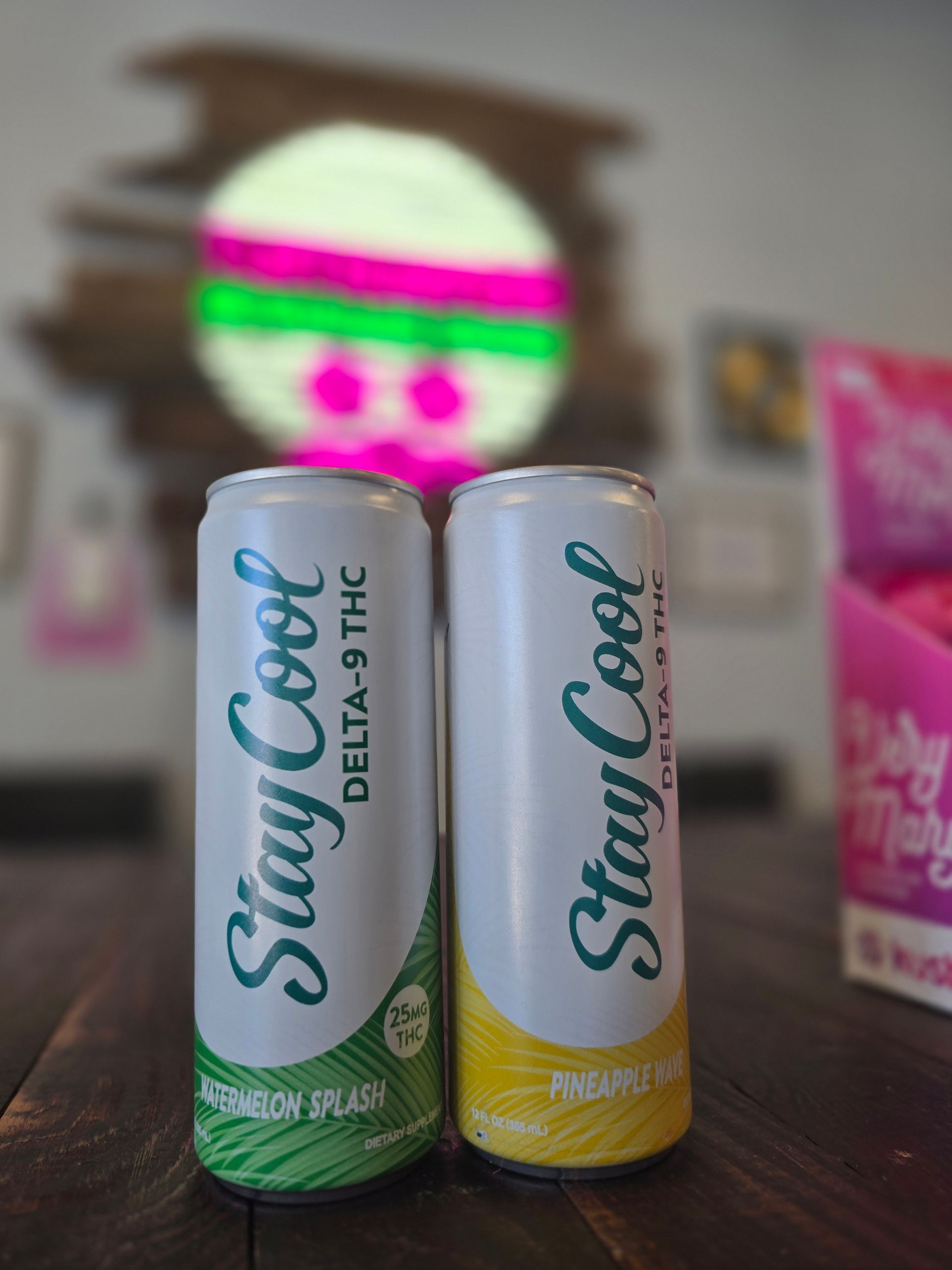 Two Stay Cool juice cans, green and yellow, on a wooden table with a blurred colorful sign in the background