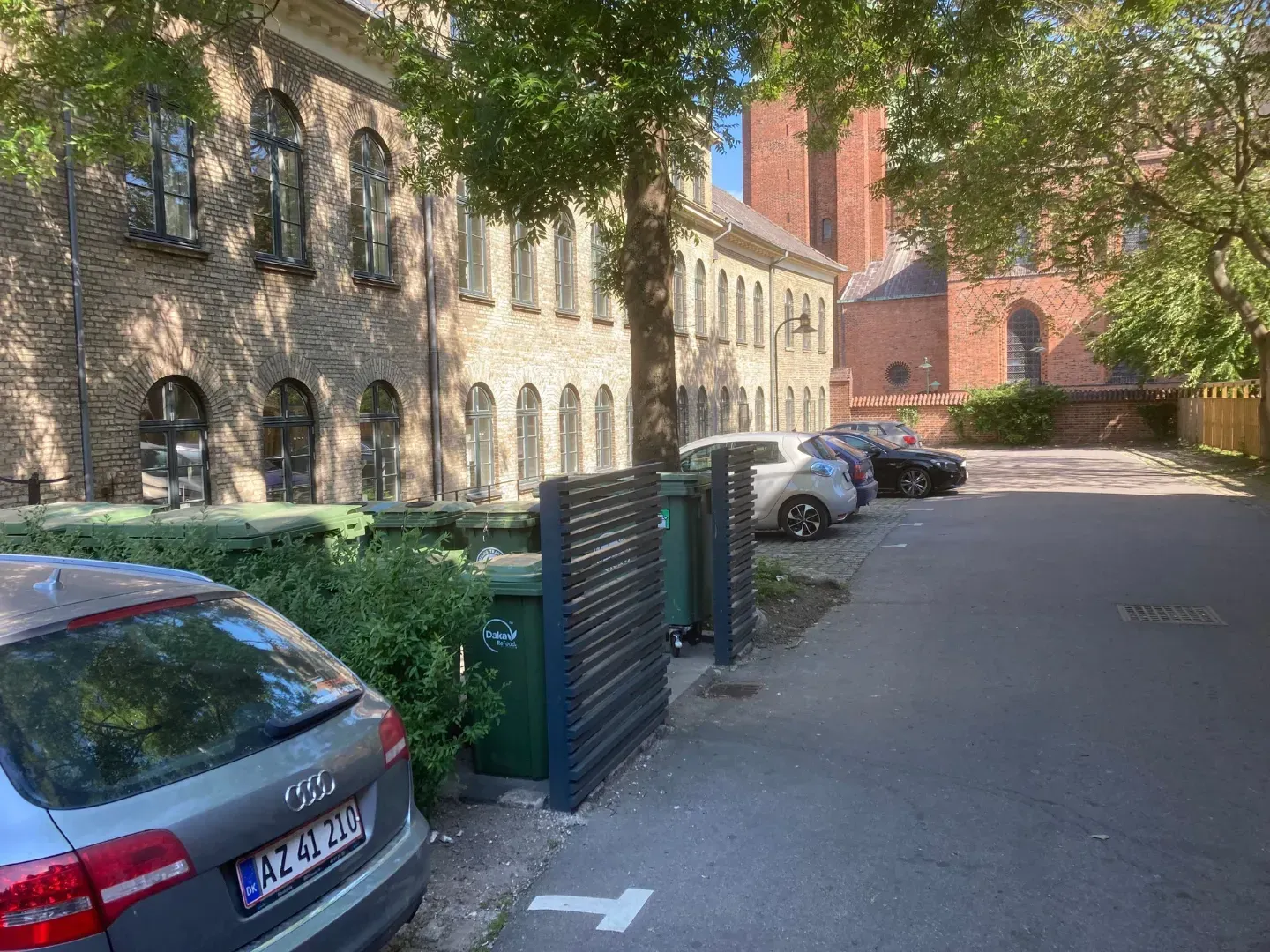 En udendørs parkeringsplads med biler parkeret nær en stenbygning og en rød murstensbygning, adskilt af et grønt hegn.