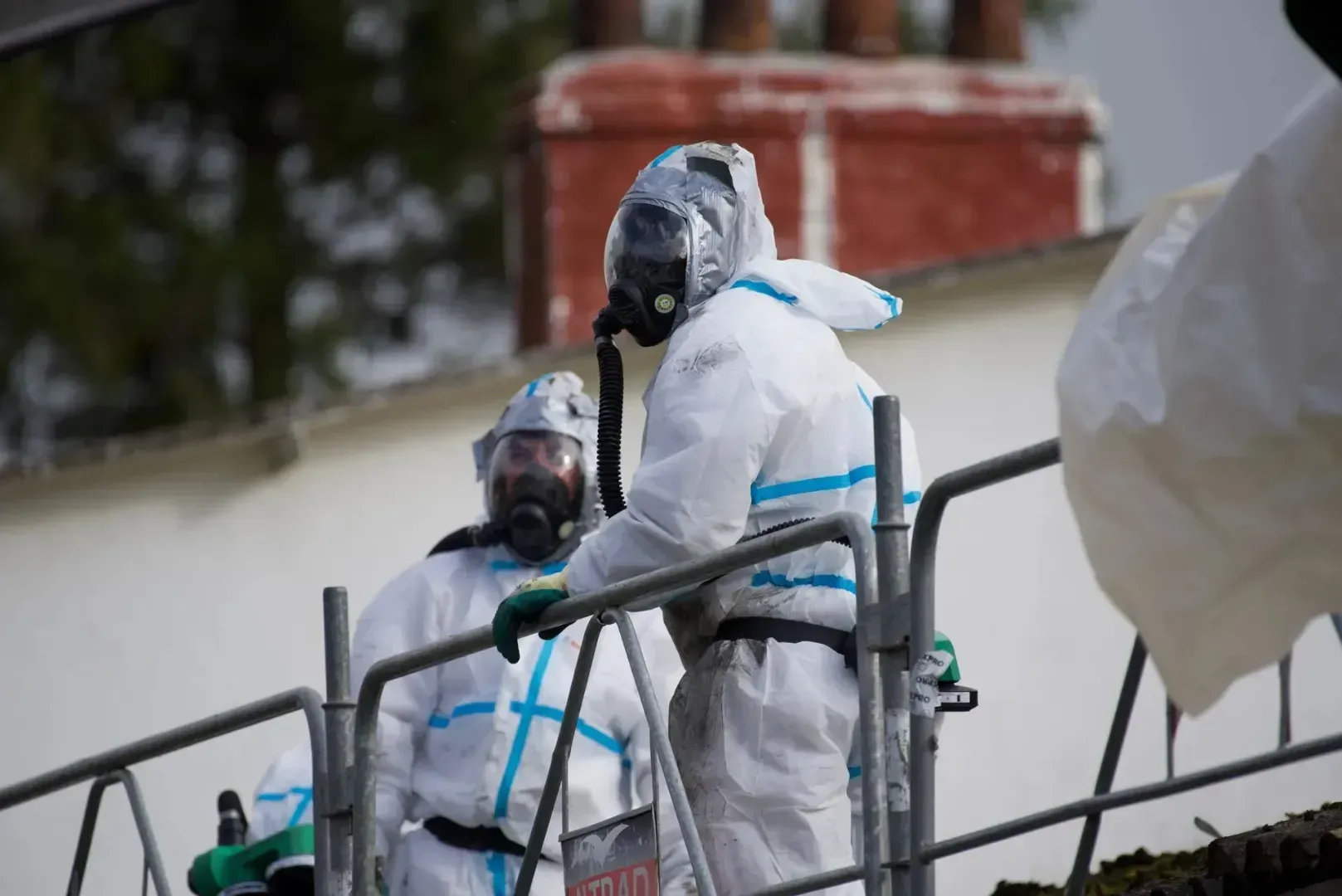 To personer i hvide dragter til farligt gods og gasmasker står på et stillads op ad en bygning med en rød skorsten.