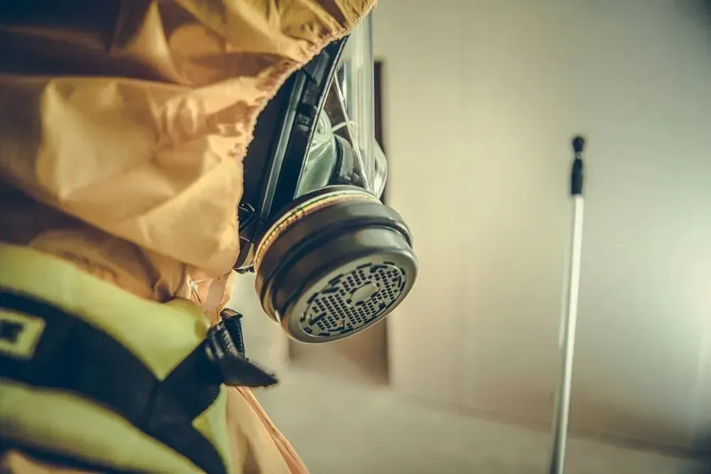 En person i en gul hazmat-dragt og åndedrætsværn står i et tomt, lyst rum med en stang i baggrunden.