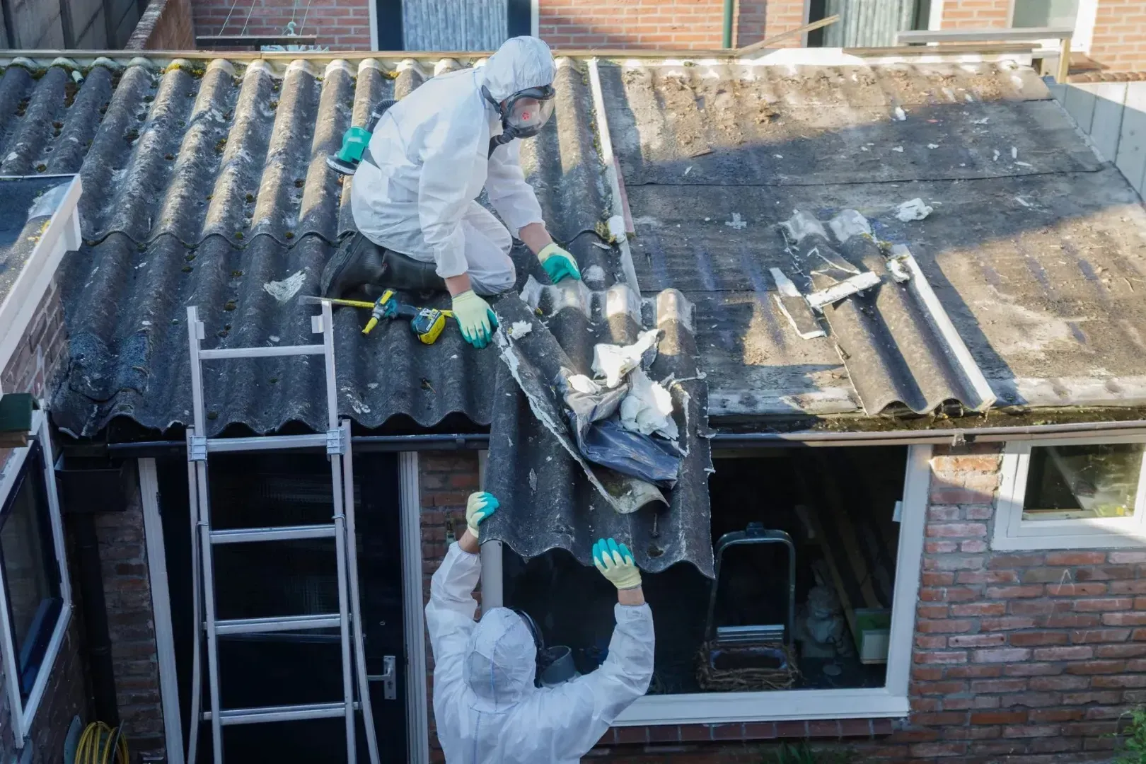 To personer i beskyttelsesdragter til håndtering af farligt gods fjerner bølgepaptag fra en beboelsesbygning.