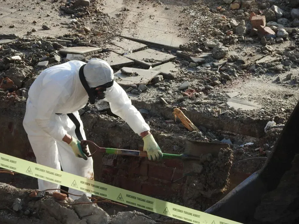 En person i en hvid hazmat-dragt og maske bruger en skovl til at rydde affald på en byggeplads bag gul advarselstape.