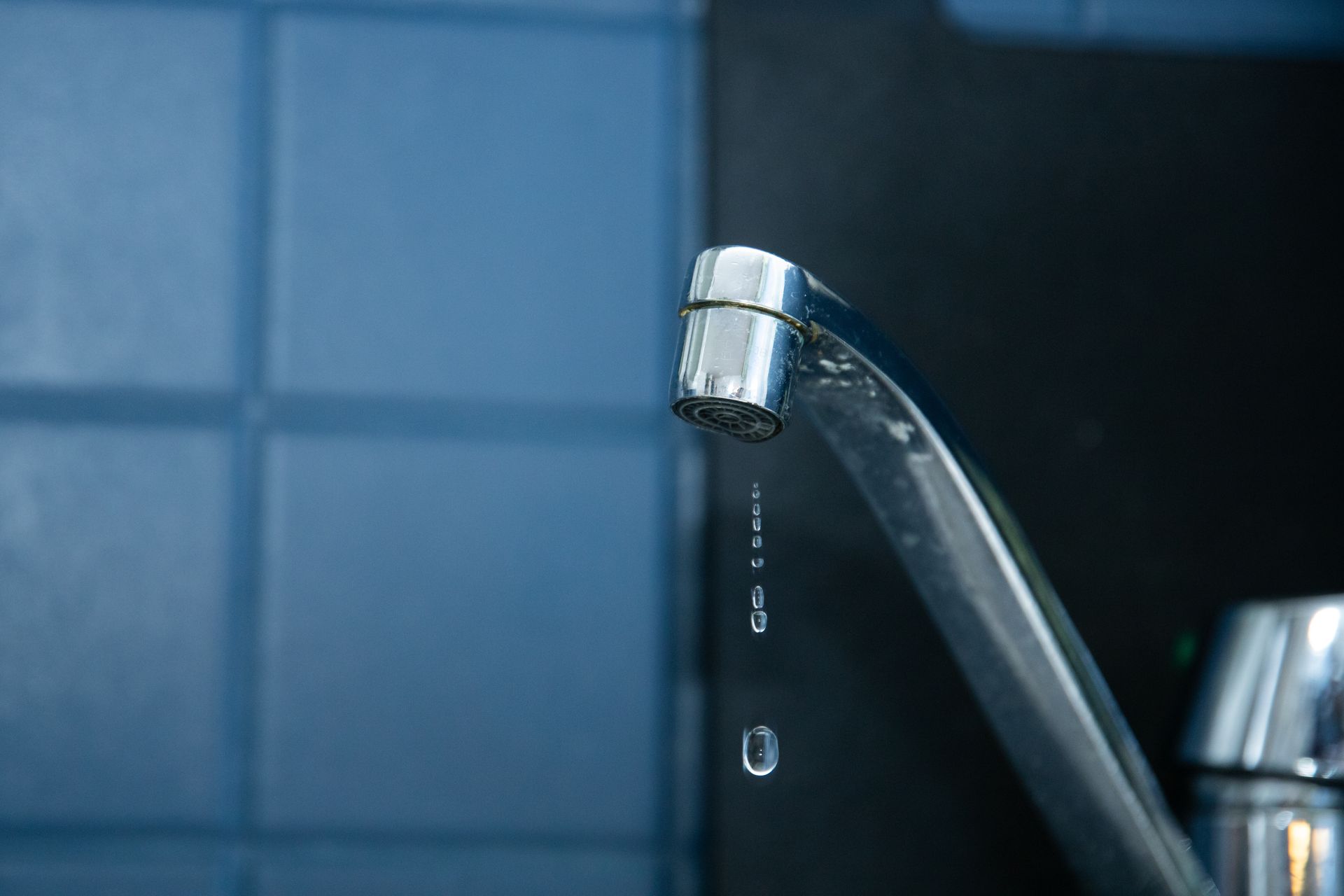 A worker in blue overalls repairs a kitchen sink faucet, with tools scattered on the countertop.