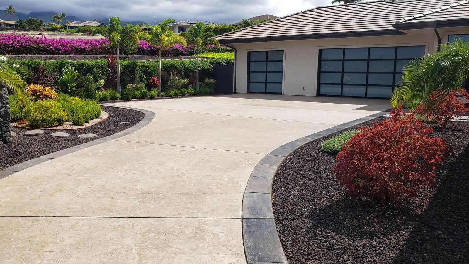 Curved concrete driveway leading to a garage, bordered by colorful landscaped shrubs and flowers.
