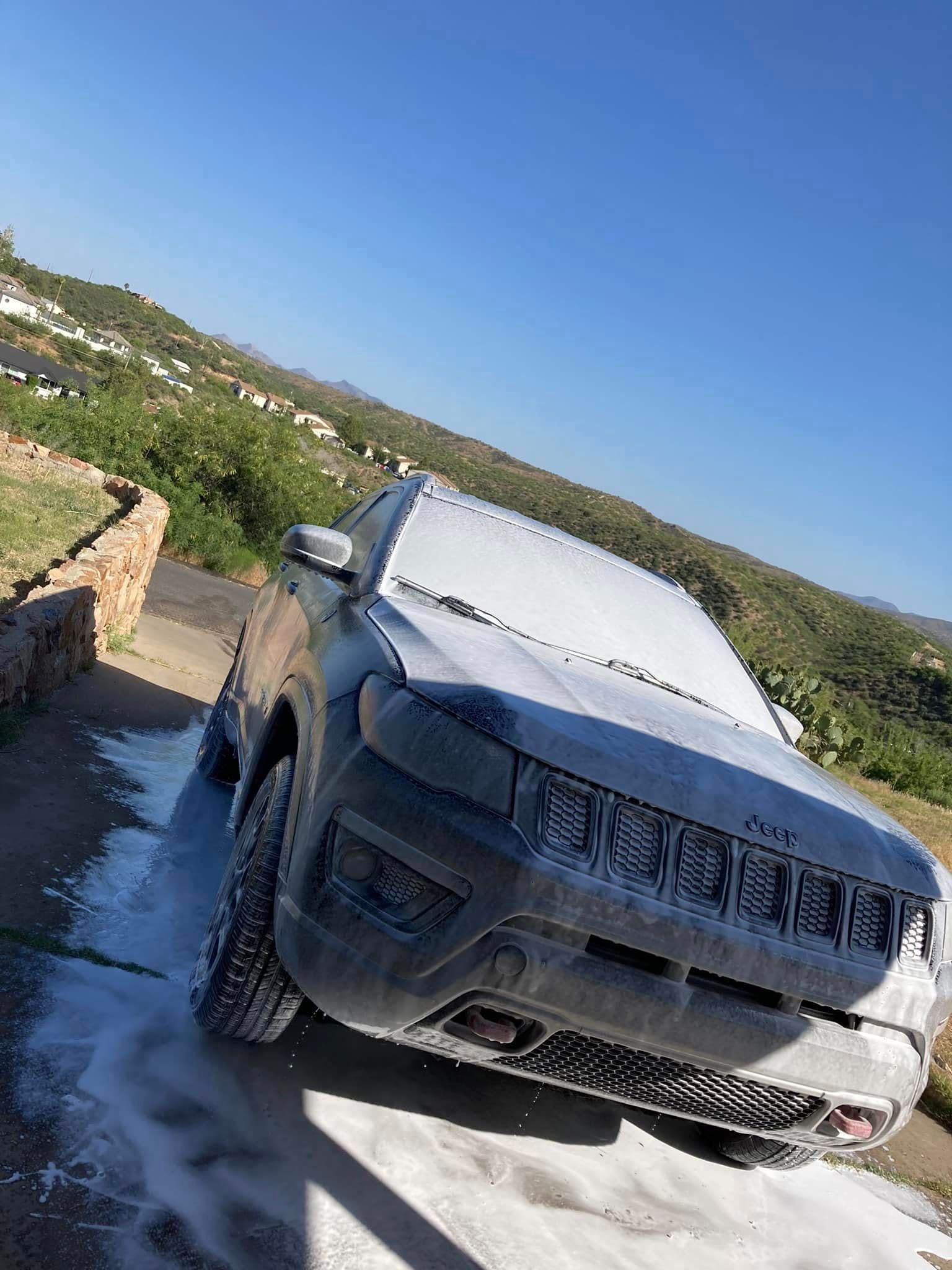 Car being washed outdoors on a sunny hillside, covered in foam with a mountain view in the background