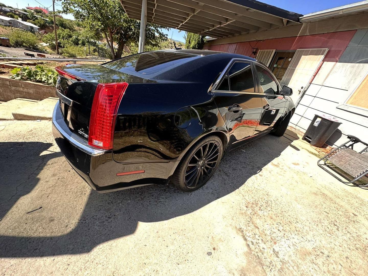 Black sedan parked in a driveway beside a house, with rear damage and a red taillight