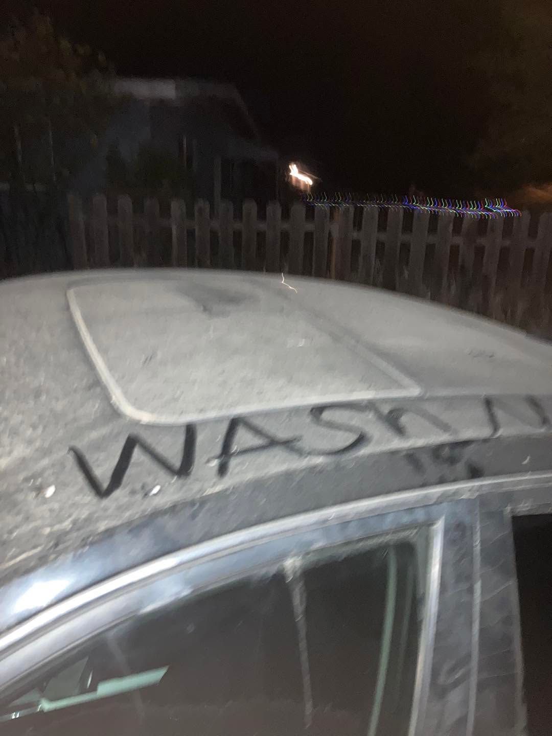 Dusty car roof with “WASH” written on it at night, beside a fence.