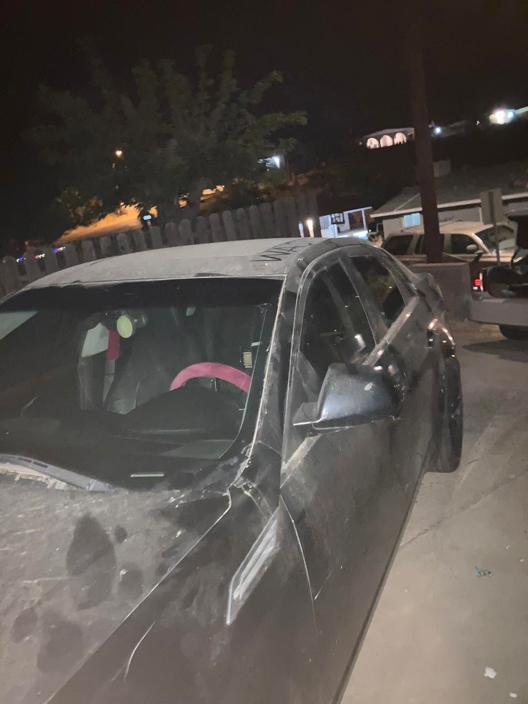 Damaged black sedan with shattered side mirror parked at night on a street by a building.