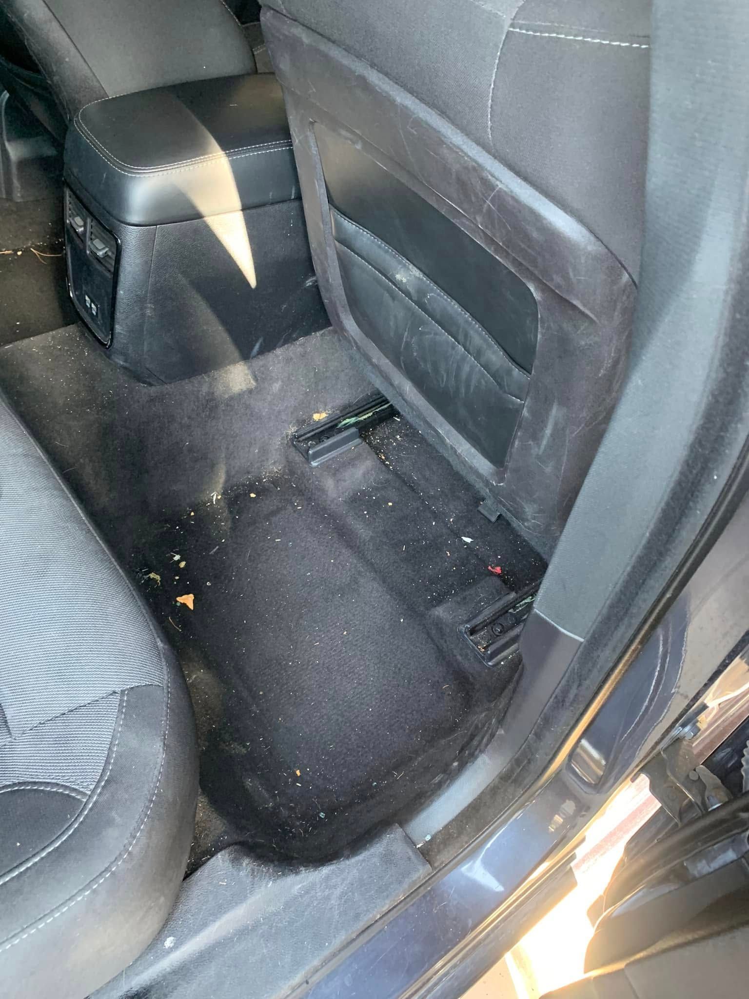 Car rear footwell with black carpet, seat back, and scattered crumbs near the center console.