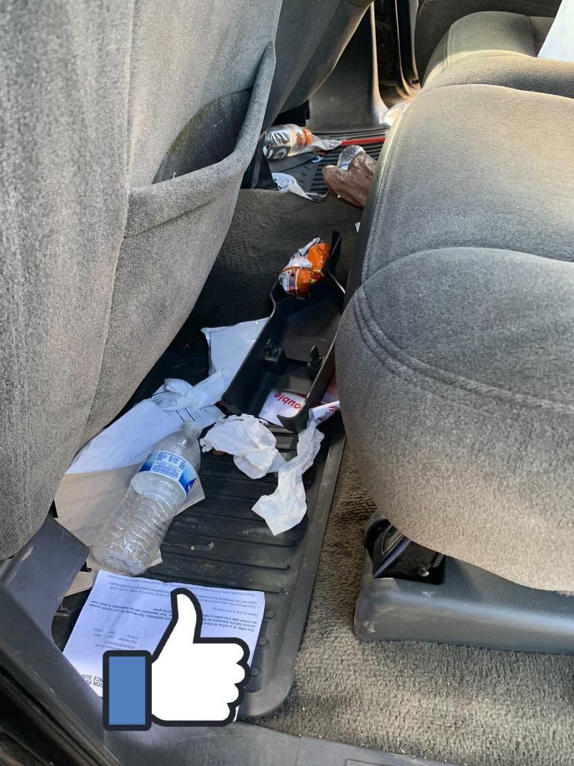 Messy car back seat floor with trash, papers, and bottles between gray seats