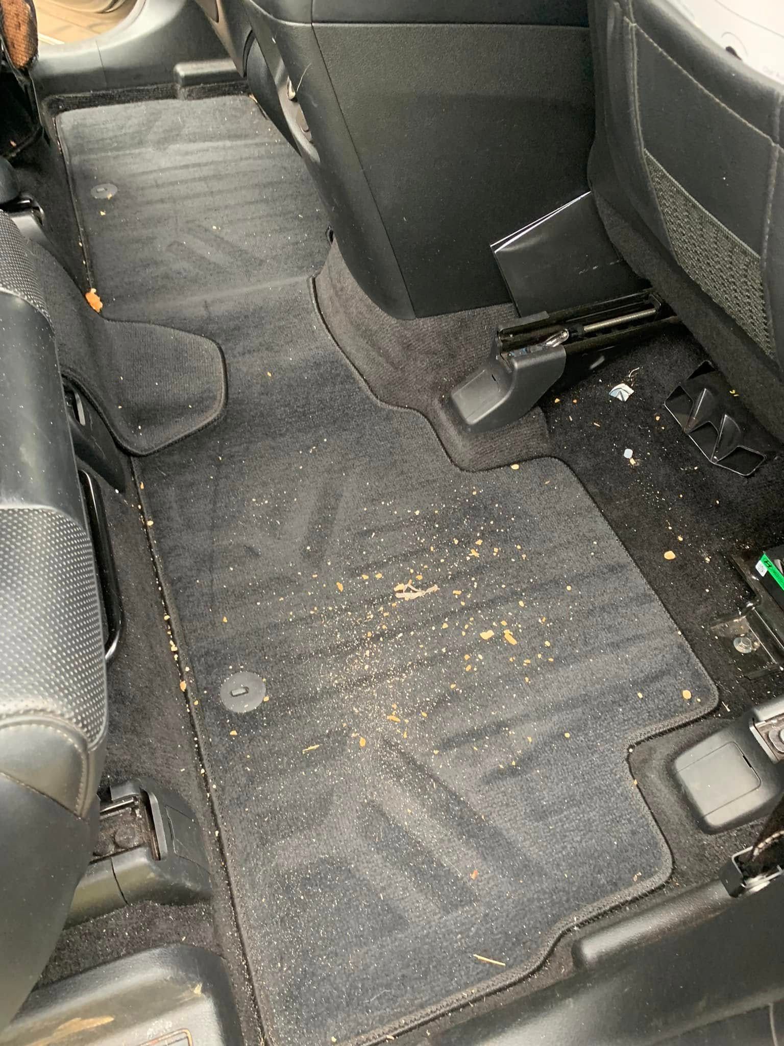 Car floor with dirty black mats, crumbs, and debris between back seats.