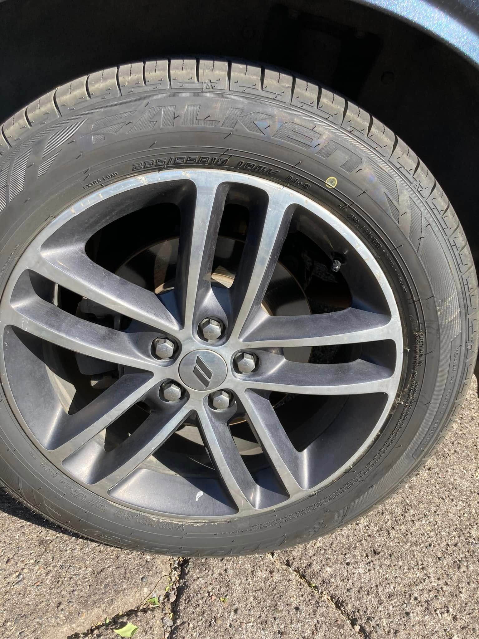 Close-up of a car tire and dark alloy wheel on pavement, with visible wear and a small scratch on the rim.