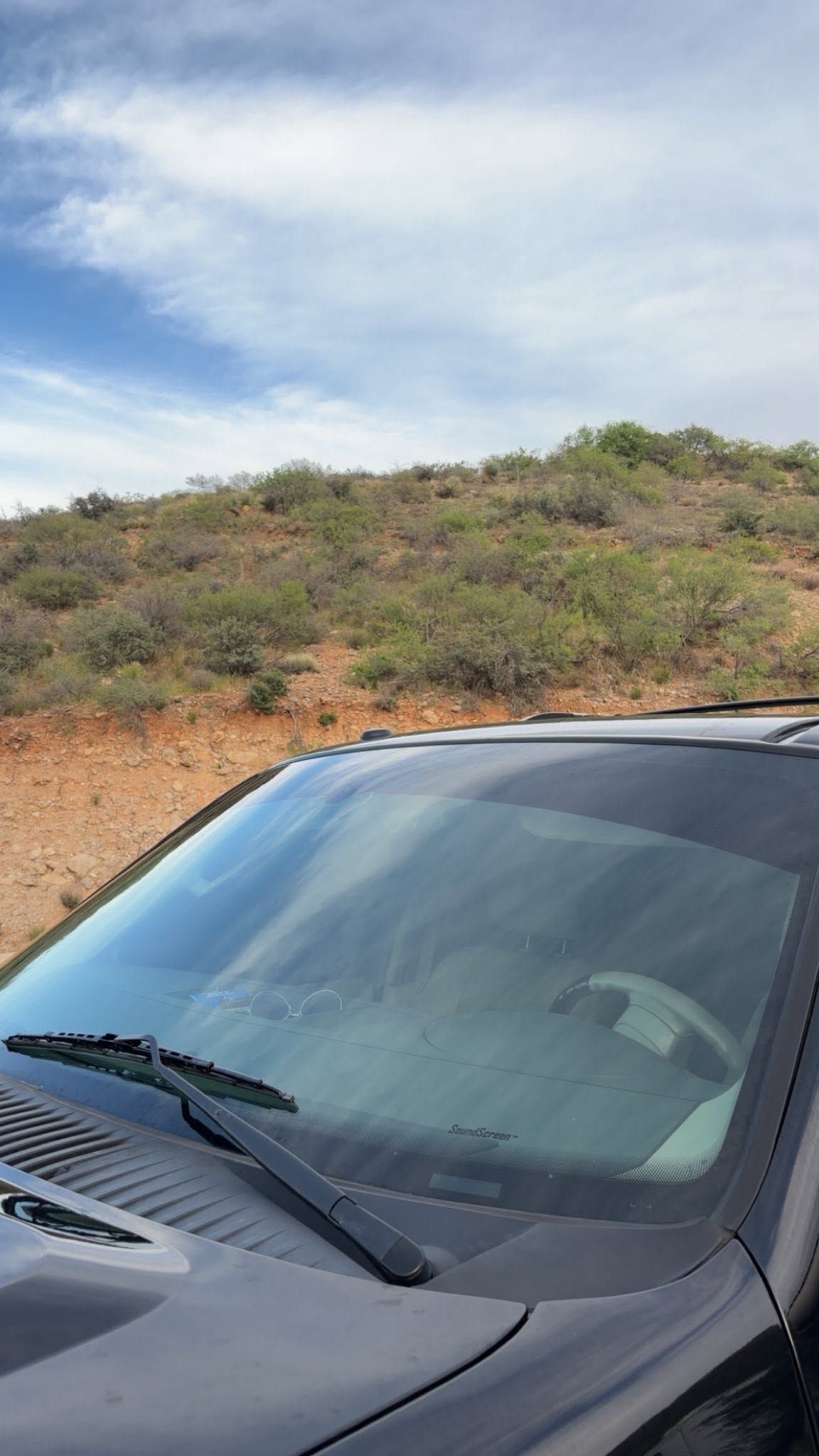 Dark gray car parked on a dirt roadside with a hillside and cloudy sky in the background