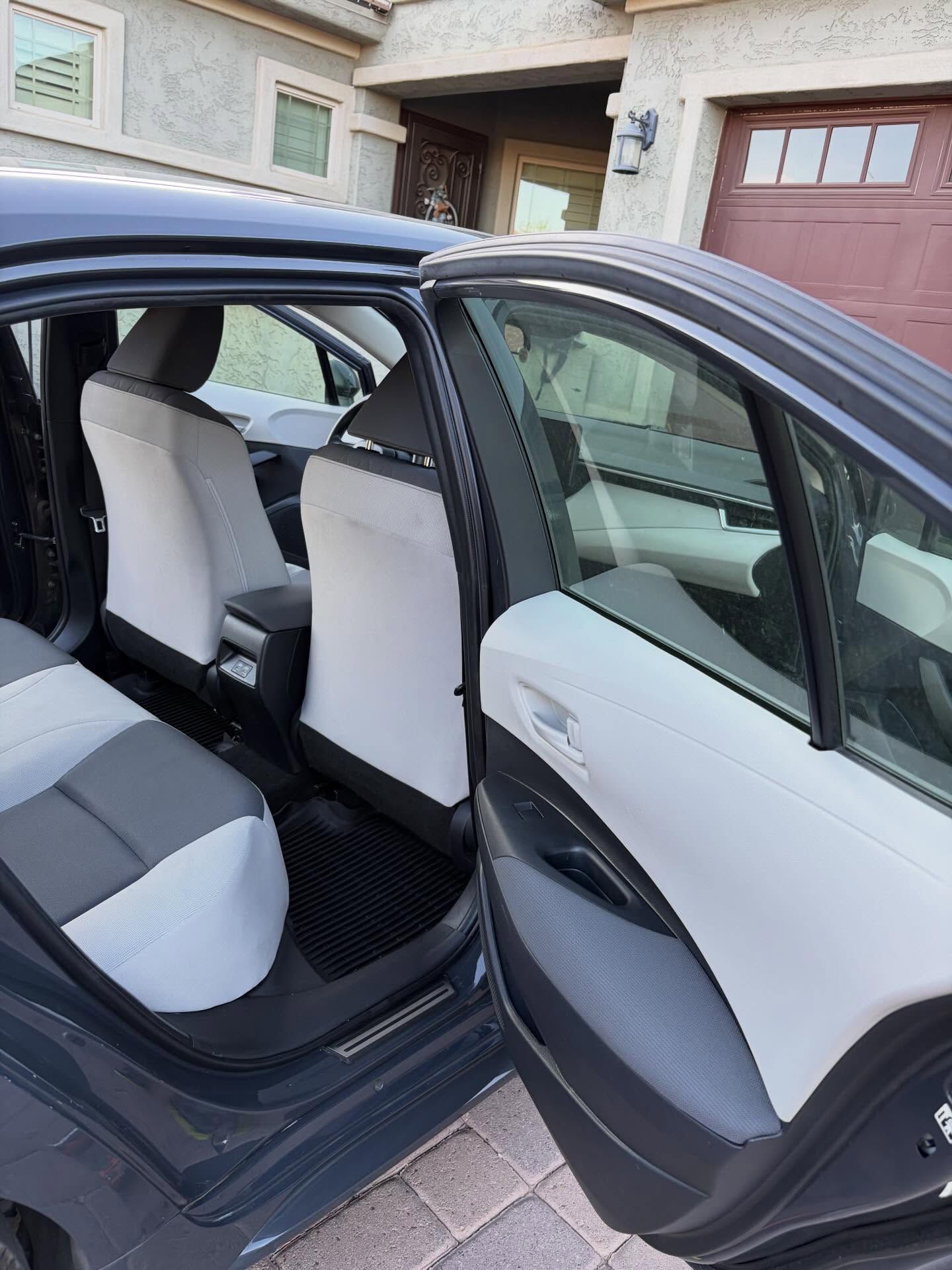 Open rear door of a dark gray car revealing white and gray interior in a driveway