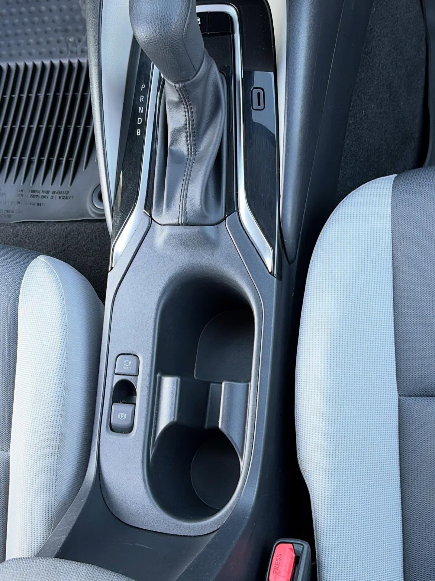 Car center console with gear shifter, cup holder, and silver trim between two gray seats