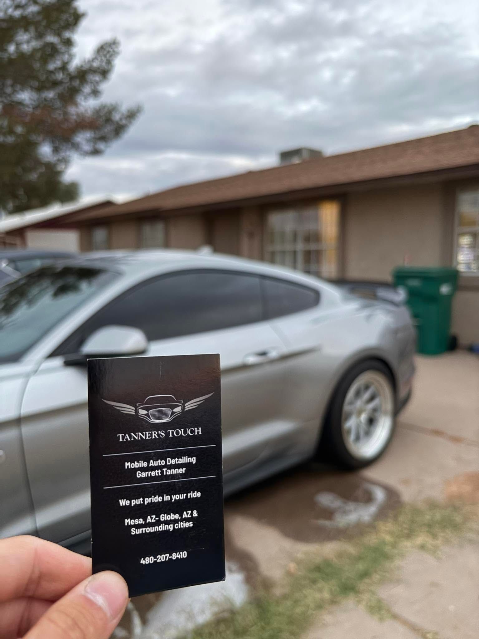 Hand holding a black card in front of a silver car parked beside a house with a green trash bin.