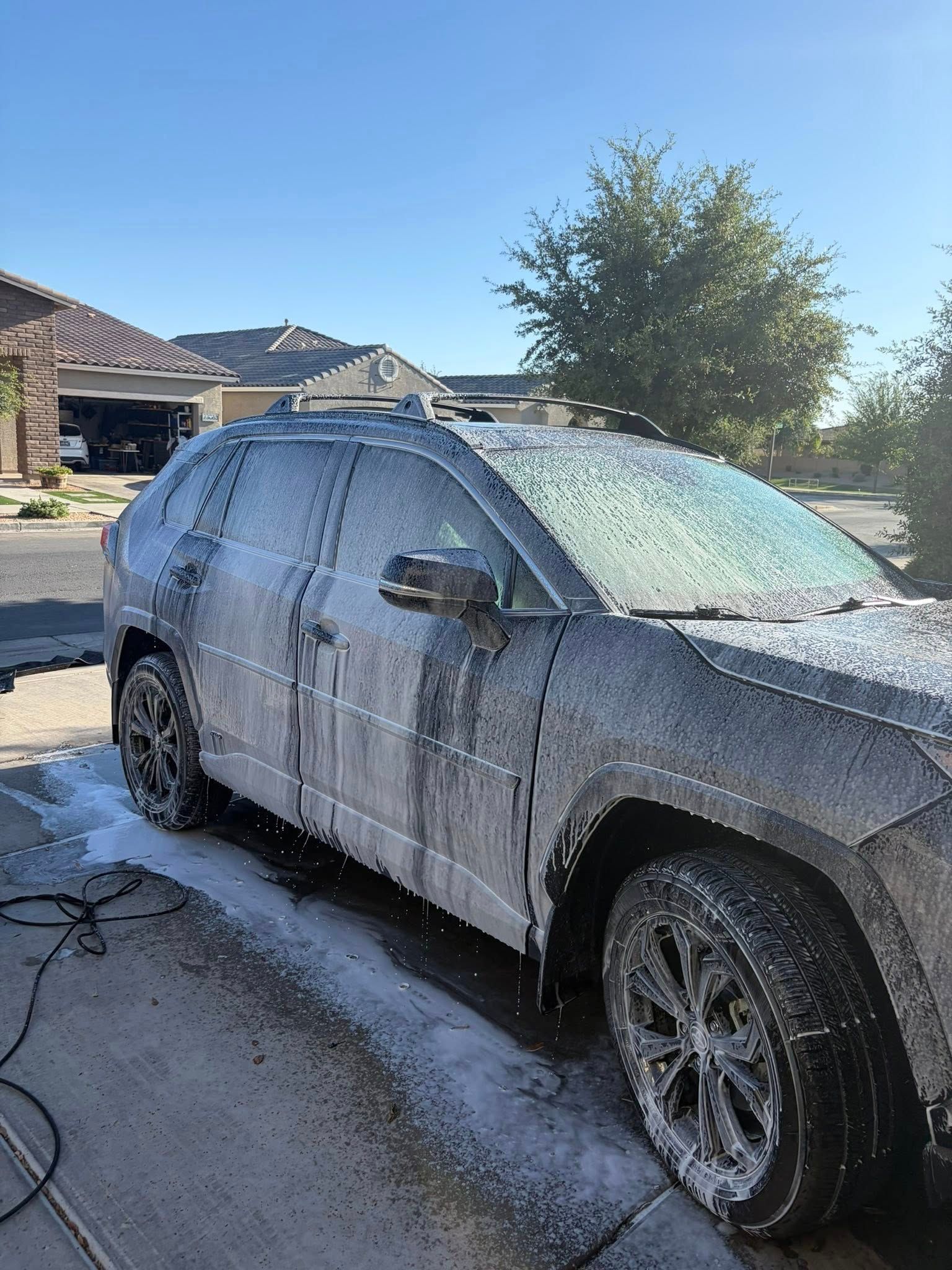 Foam-covered gray SUV parked in a driveway being washed in bright sunlight