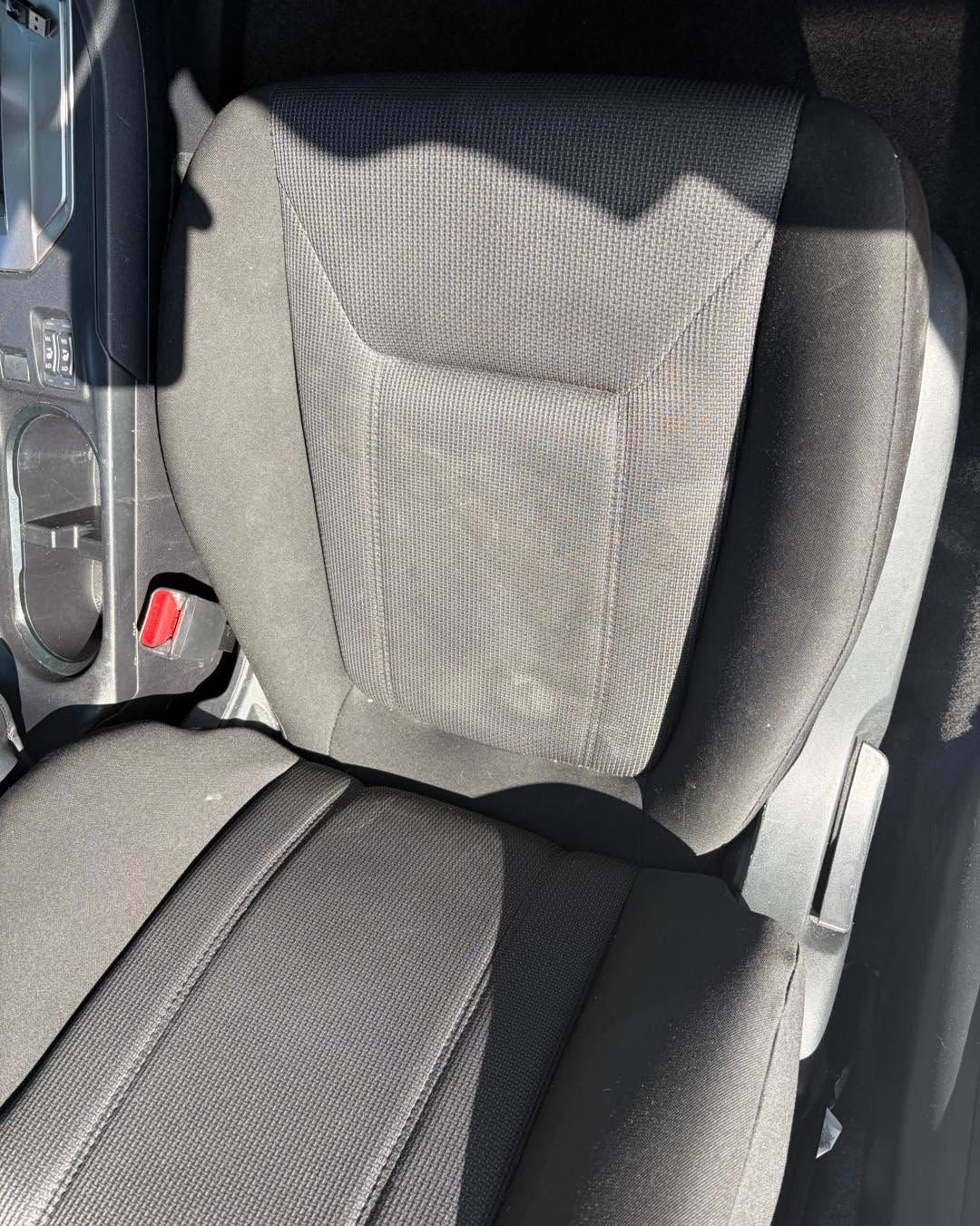 Gray car seat with a wrinkled light cover in bright sunlight, beside a black seat and red seatbelt latch