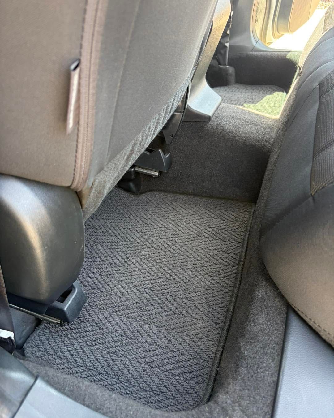 Carpeted floor area in a car between two seats, with gray upholstery and sunlight visible in back.