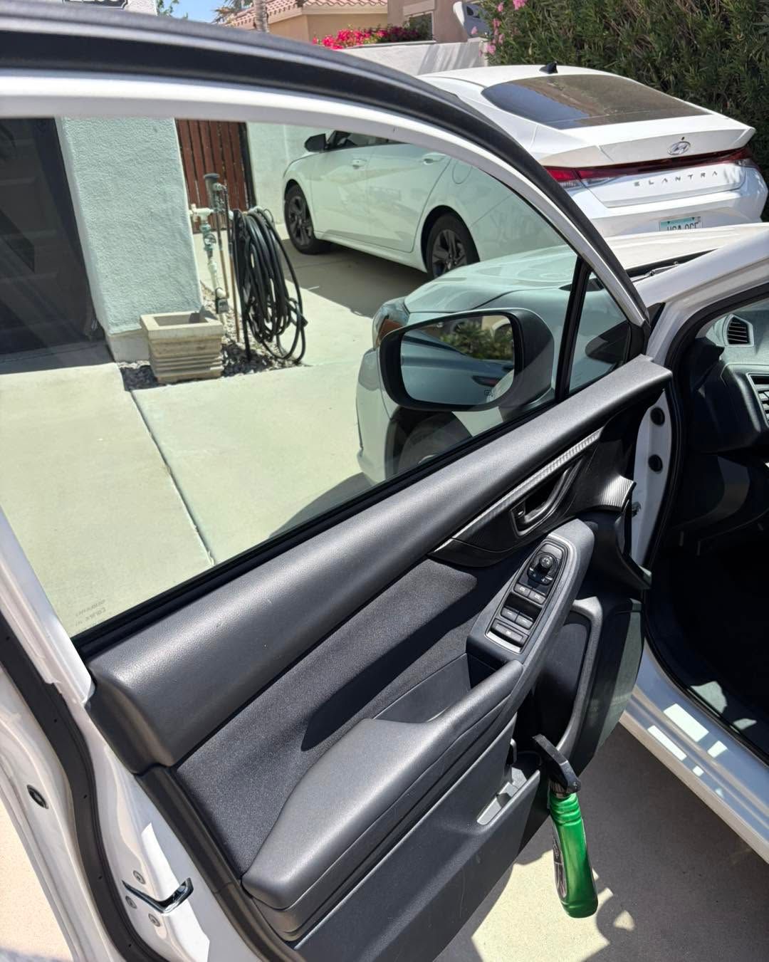 Open car door showing black interior, green sanitizer bottle, and a parked white car outside.