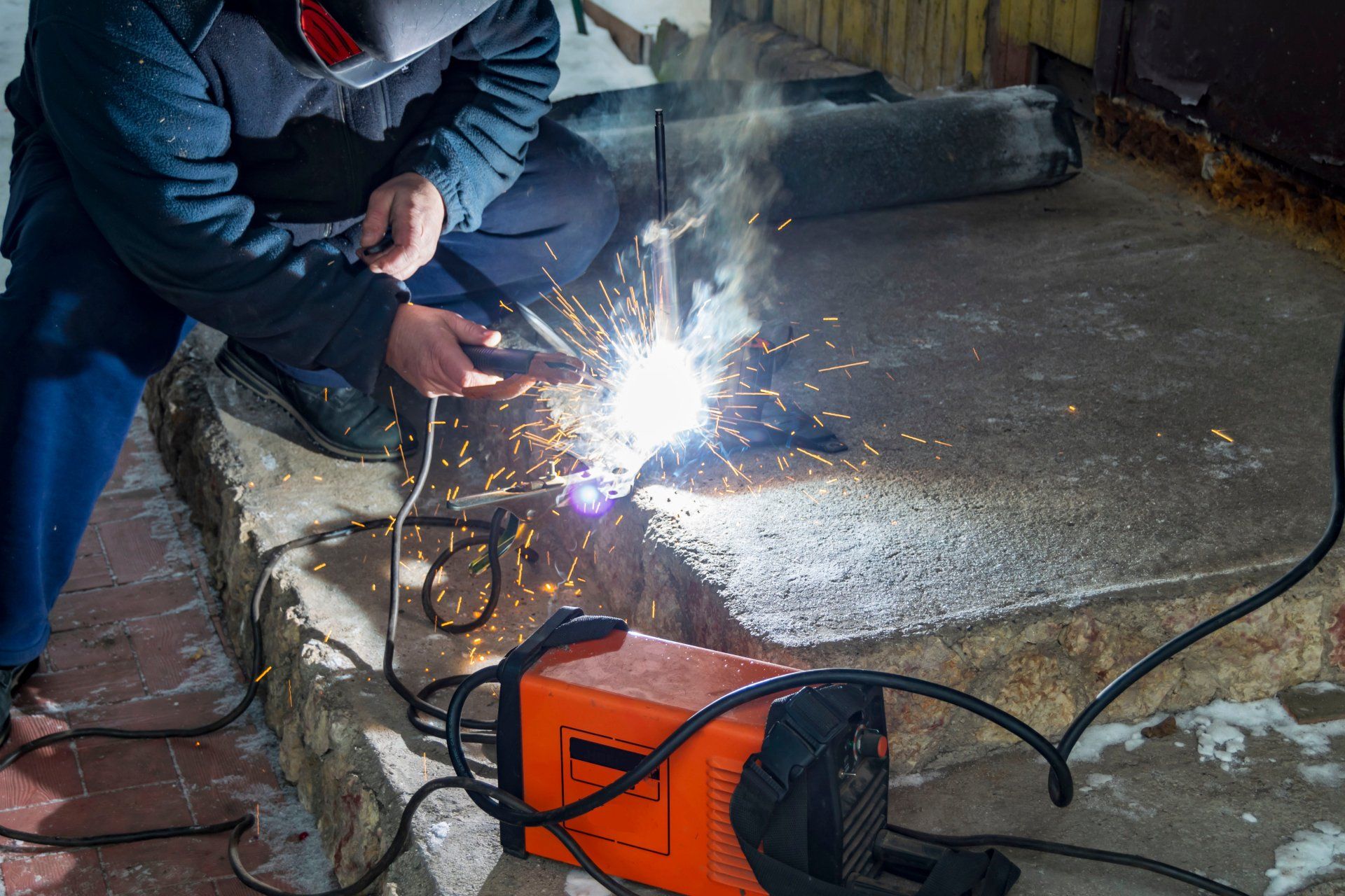 Soudeur en vêtements sombres travaillant du métal avec des étincelles, soudeur orange sur du béton, en intérieur.