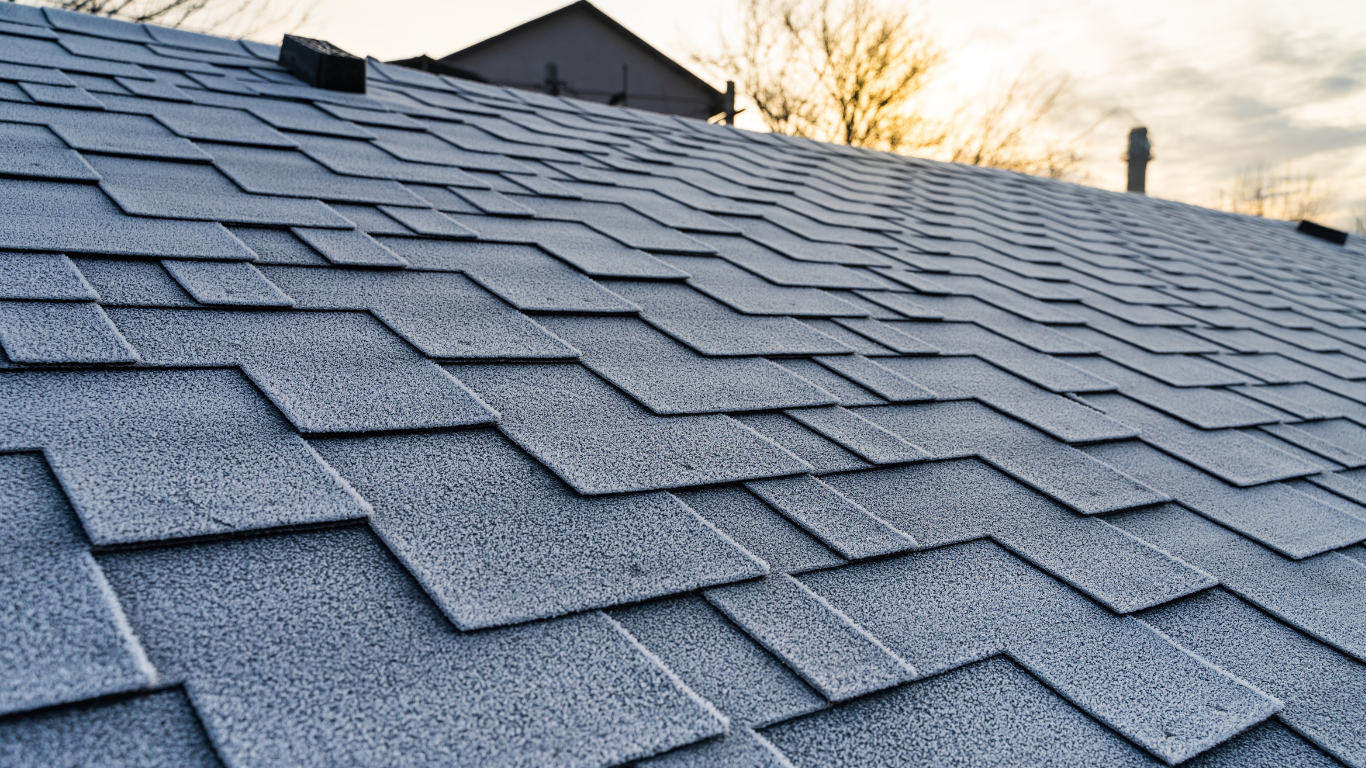 Close-up of a gray shingled roof with frost, angled perspective. The sun is setting in the background.